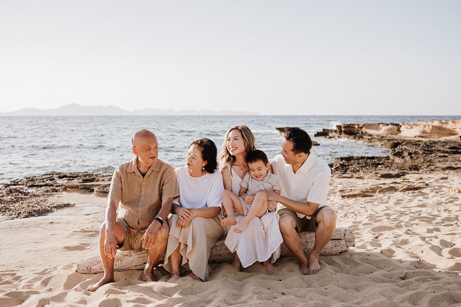 Multigenerational family smiling and laughing together on a sandy beach with ocean views on a sunny day.