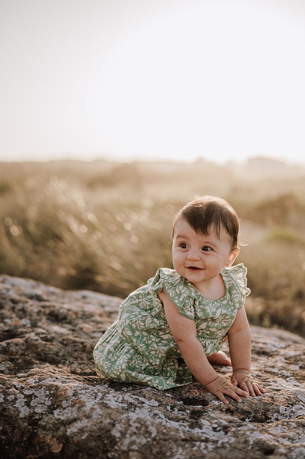 A baby girl in a green floral romper crawls on a rock outdoors with golden sunlit grass in the background.