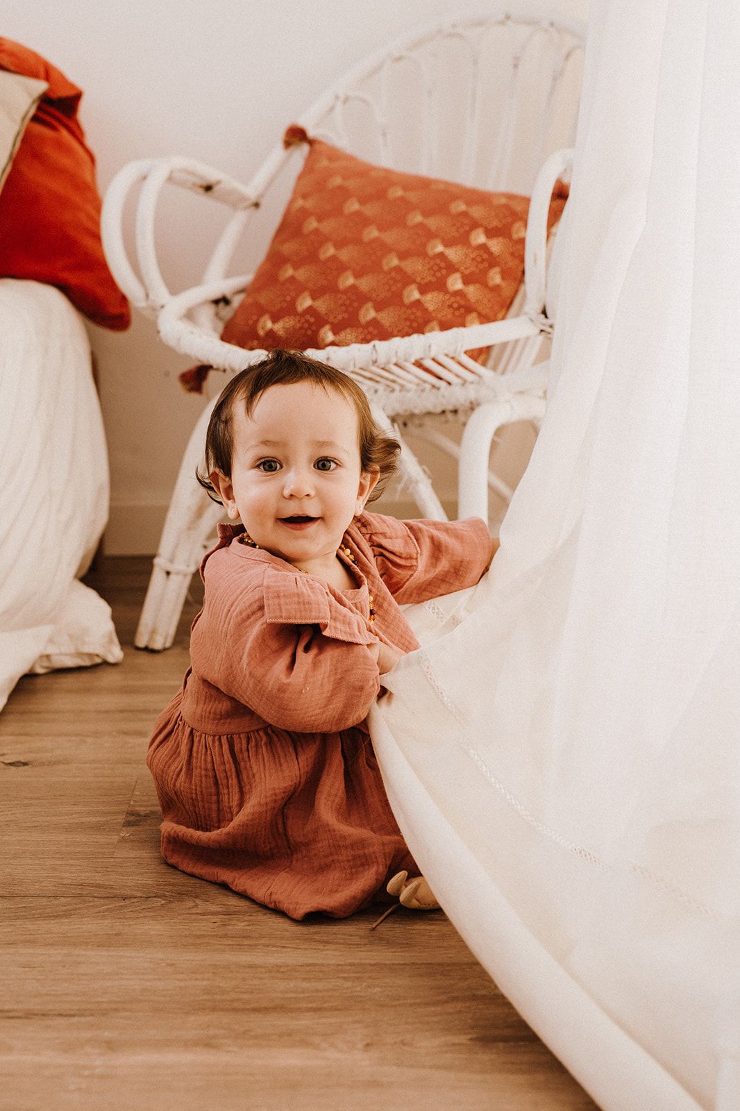 Adorable toddler girl in a rust-colored dress sitting on hardwood floor next to a white rattan chair.