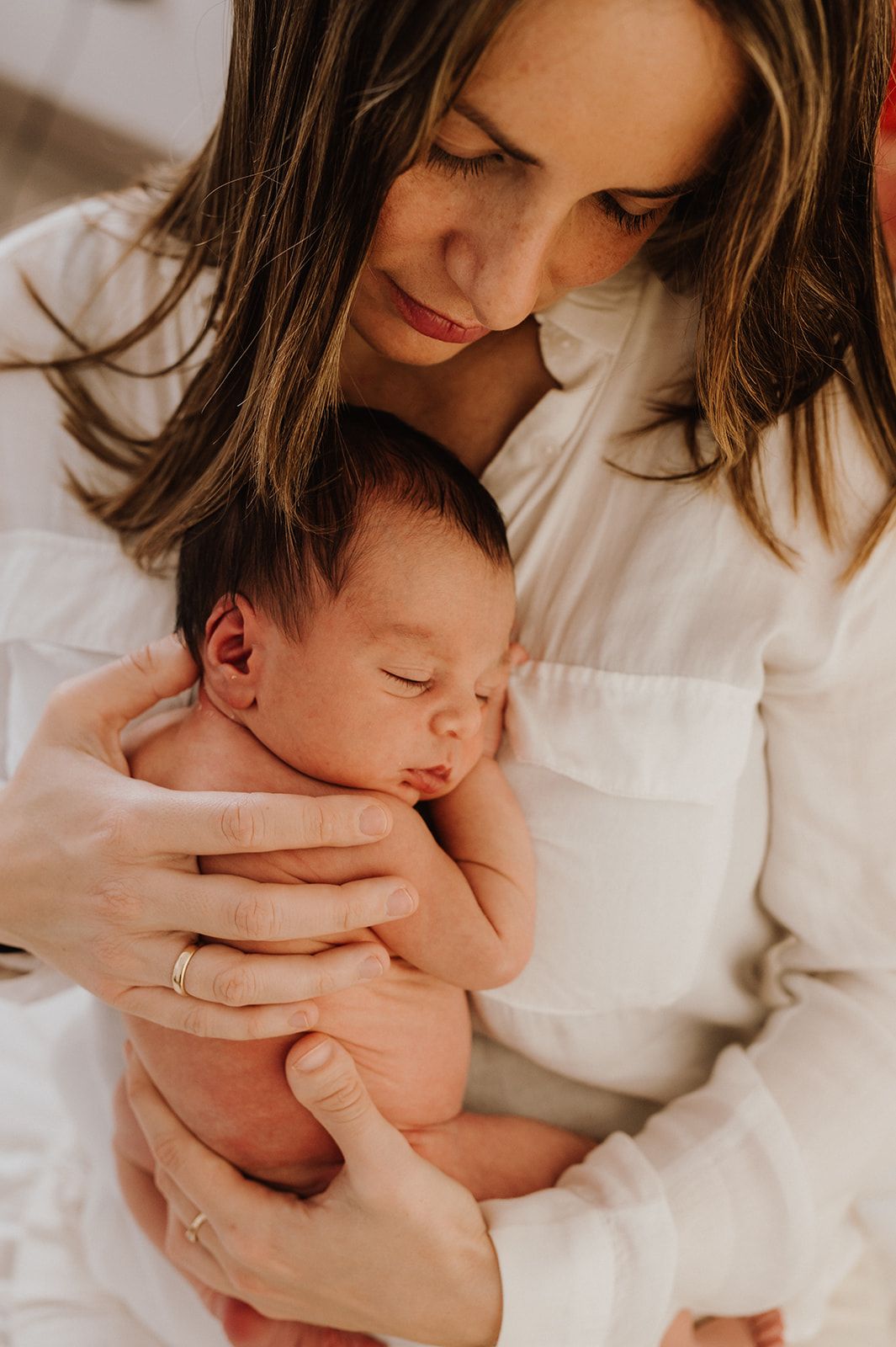 Mother in white shirt tenderly holding sleeping newborn baby with eyes closed in her arms.