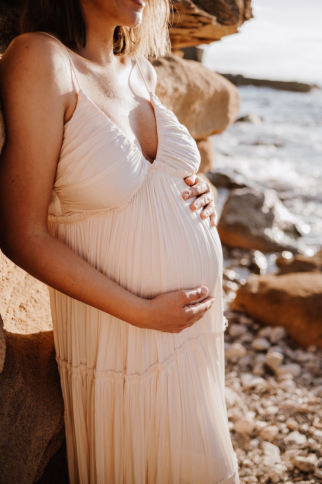 Pregnant woman in beige dress cradling baby bump beside rocky ocean shore in warm golden light.