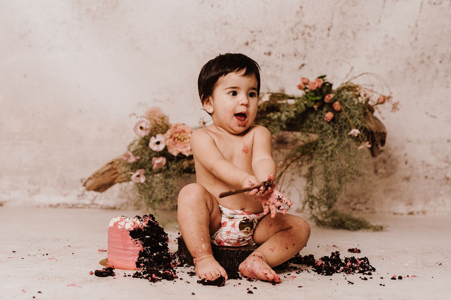 Baby's first birthday cake smash session with pink floral backdrop, chocolate cake, and adorable messy toddler.