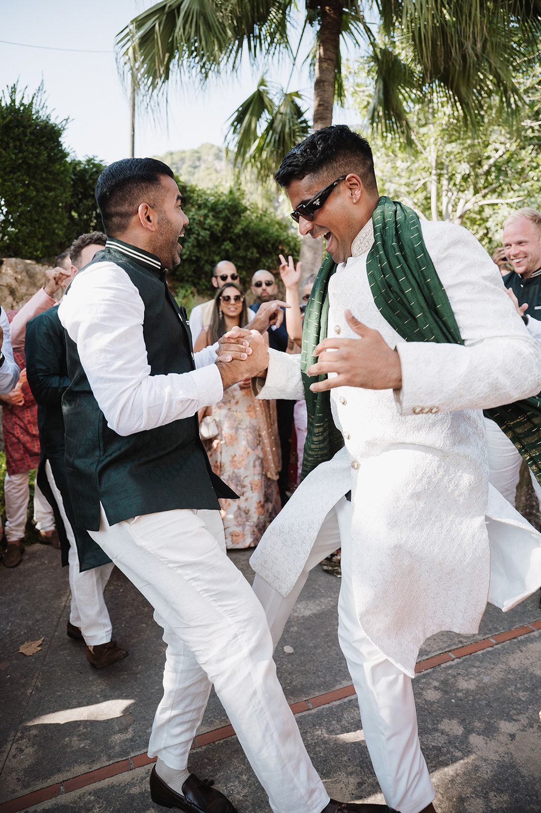 Two men in white Indian wedding attire dance joyfully outdoors at a traditional South Asian wedding celebration.