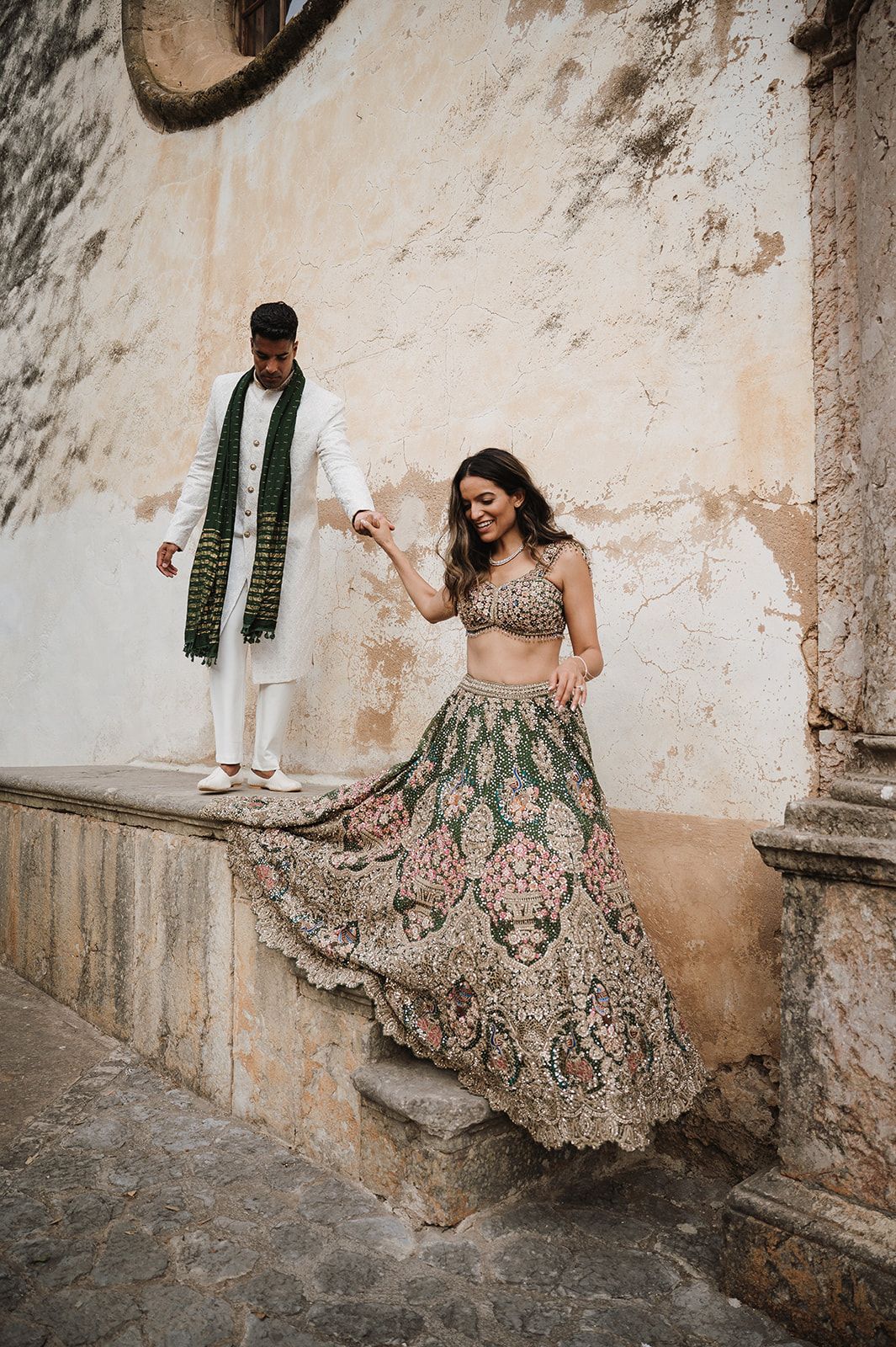 Groom in white sherwani helps bride in ornate green lehenga descend stone steps at historic venue.