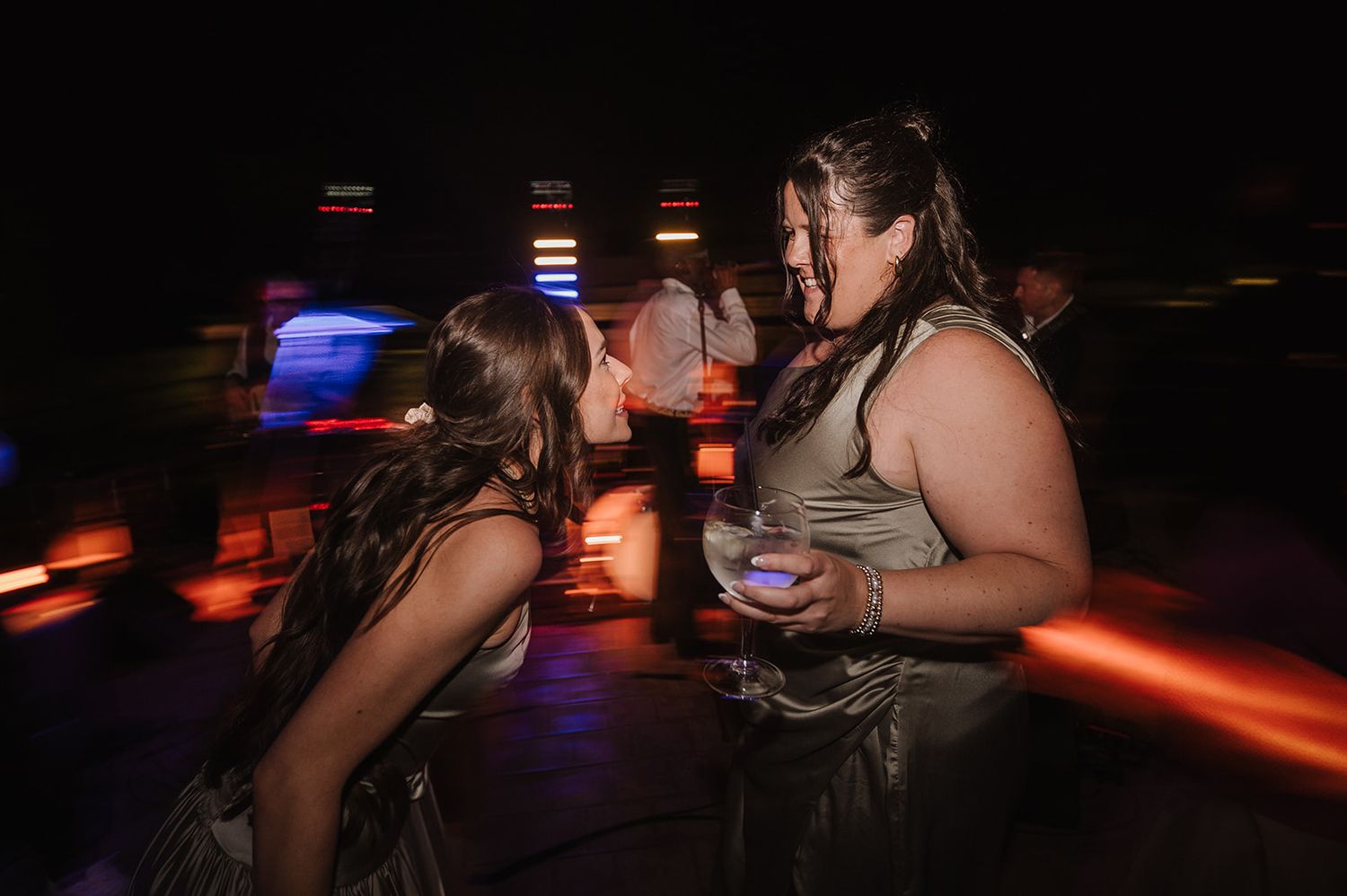 Two women laugh and chat together on a vibrant, colorful nighttime wedding dance floor.