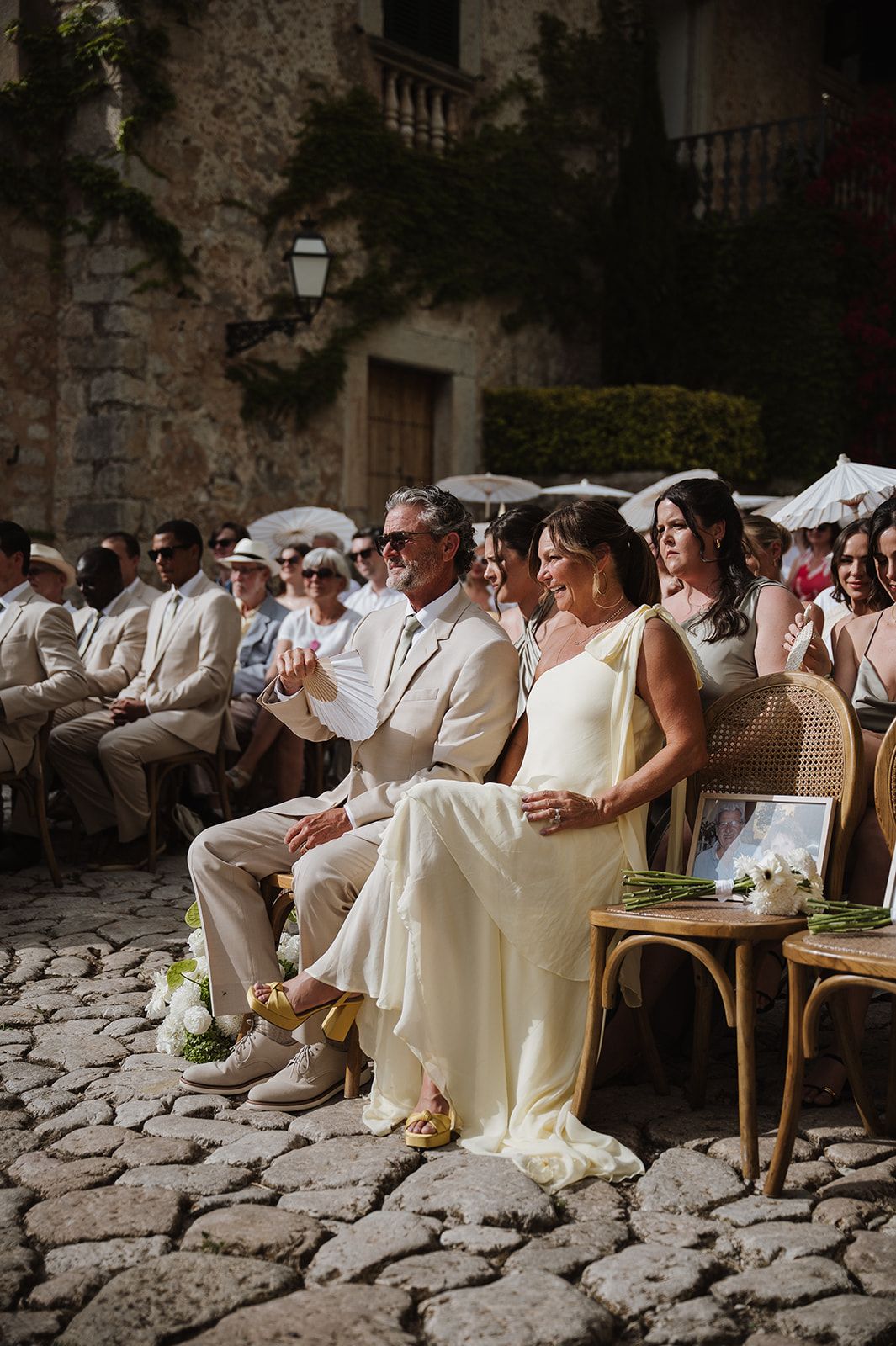 Elegant wedding guests seated outdoors on cobblestones at a rustic European venue during a sunny ceremony.