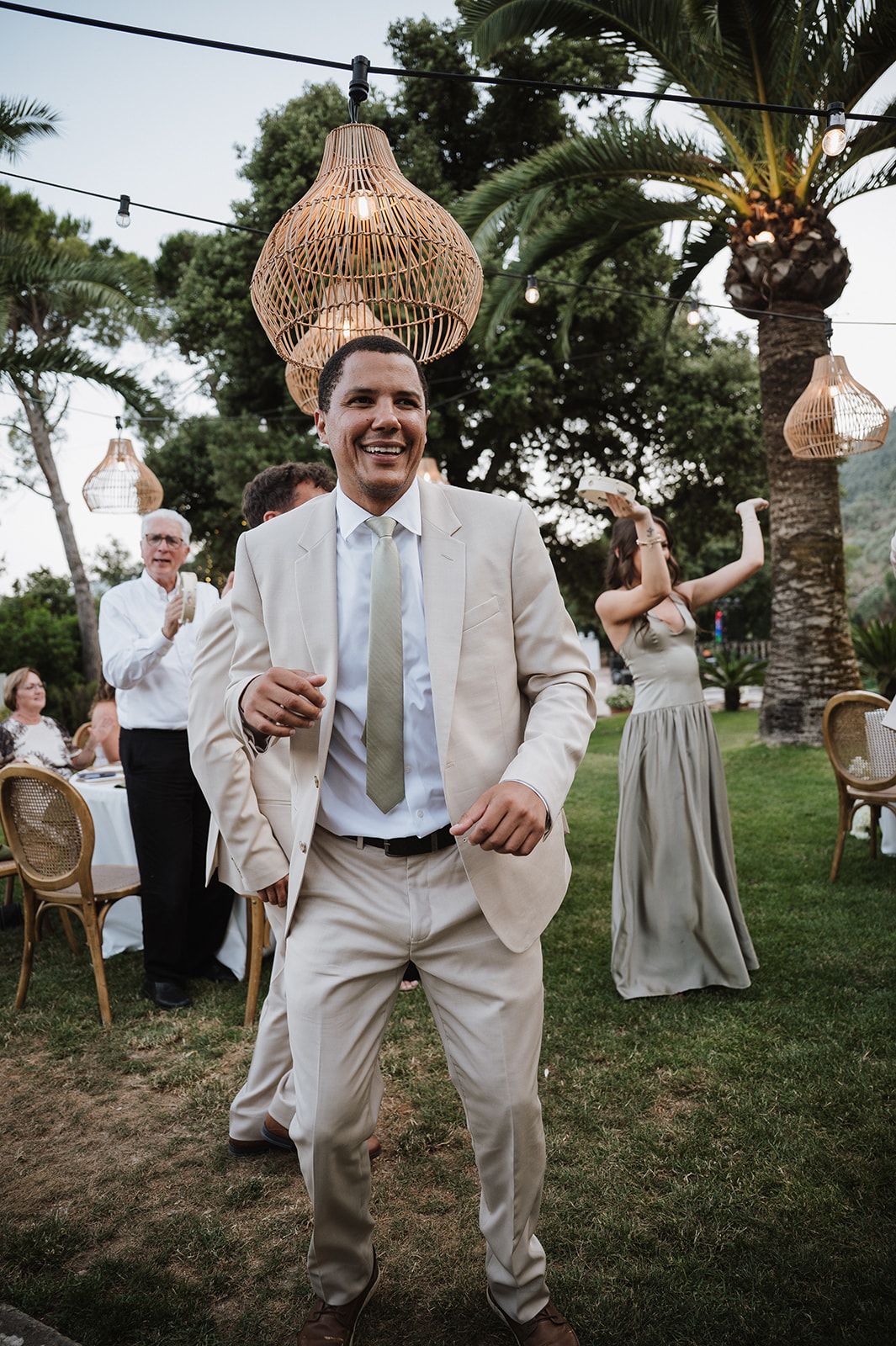 Joyful groom in beige suit dancing at outdoor wedding reception under rattan pendant lights and palm trees.
