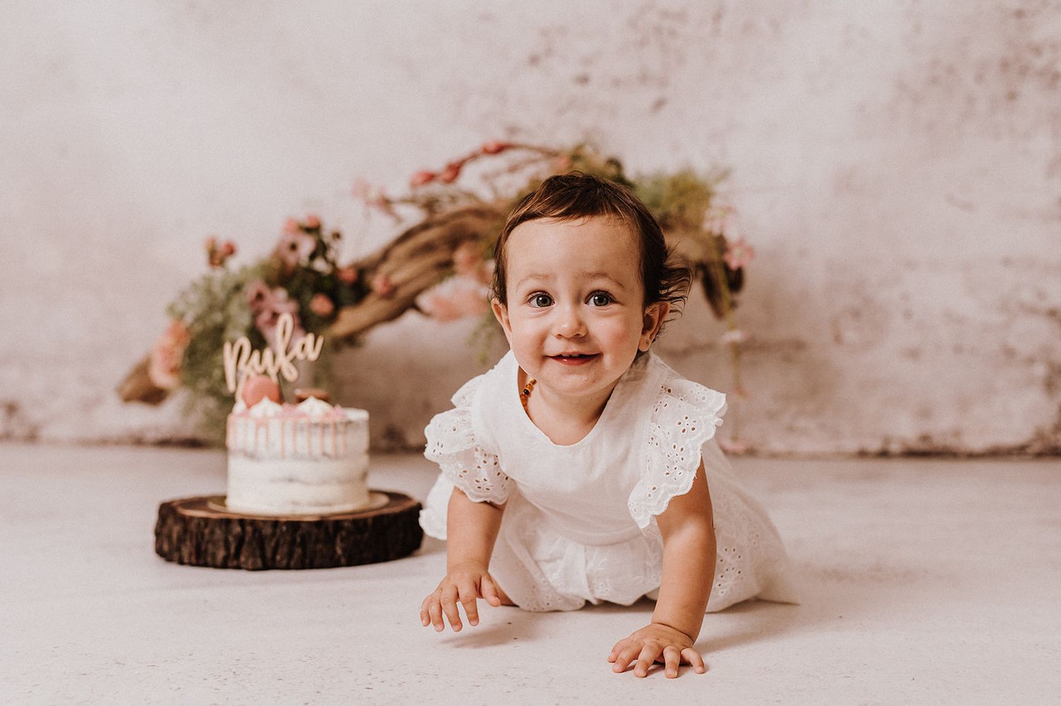 Baby girl in white dress crawls toward a small birthday cake on a wood slice during a first birthday cake smash photo session.