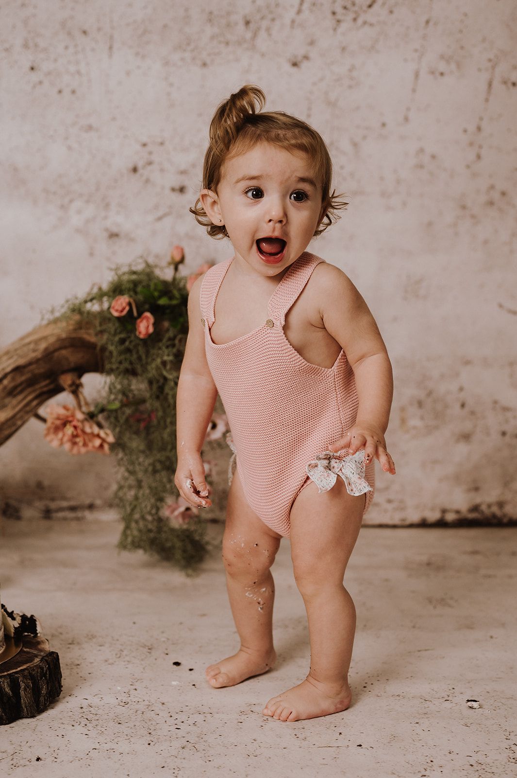 Toddler girl in pink ribbed romper standing barefoot with mouth open, posed in front of floral backdrop.