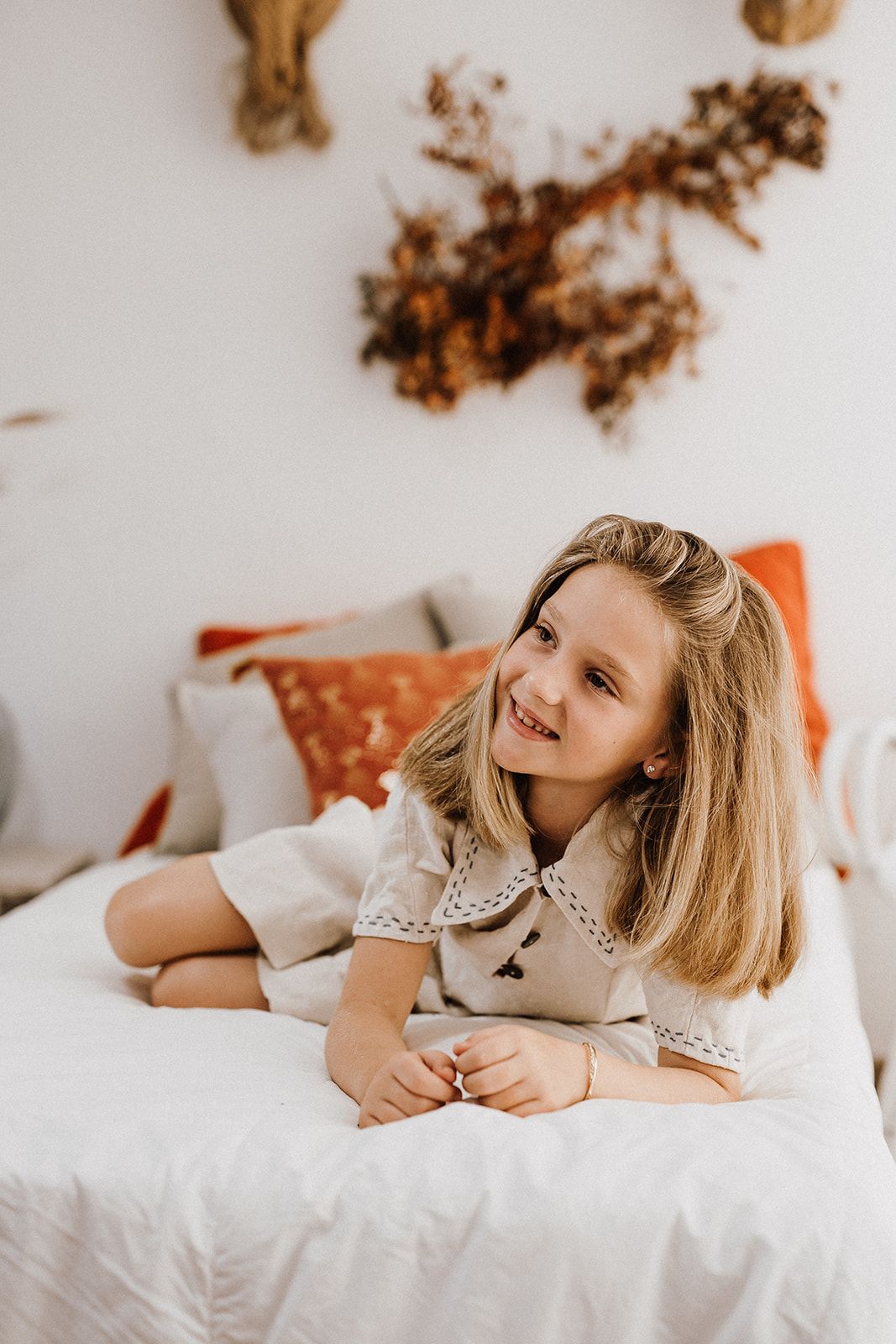 Smiling blonde girl in white dress lying on white bed with orange pillows and dried floral wall decor.