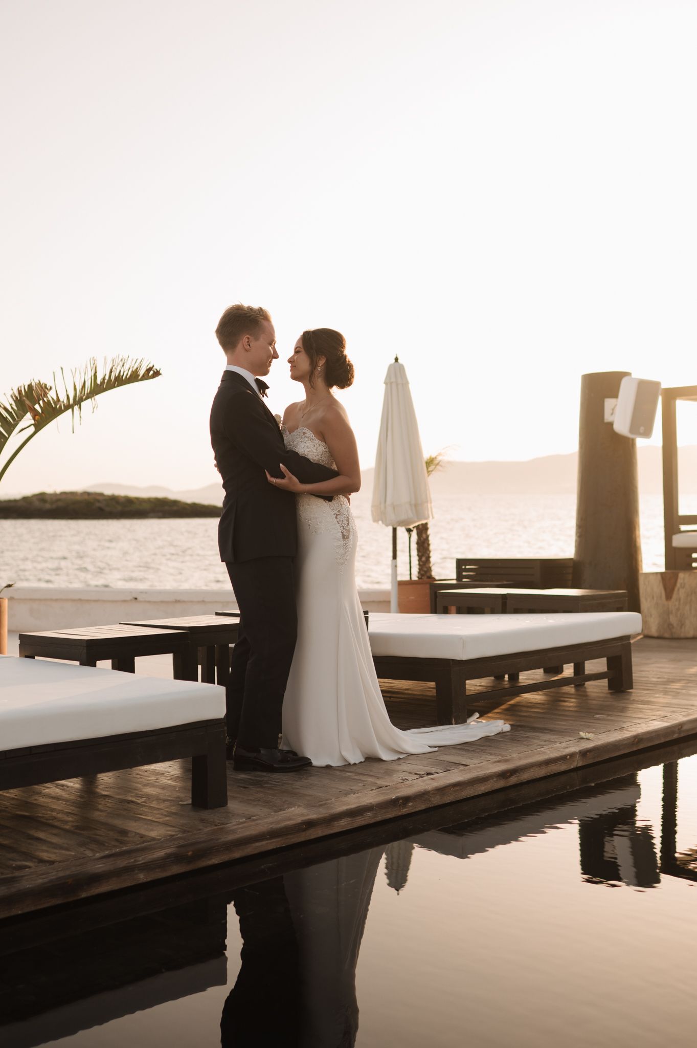 Bride and groom embracing on a waterfront dock at golden sunset during their wedding day.