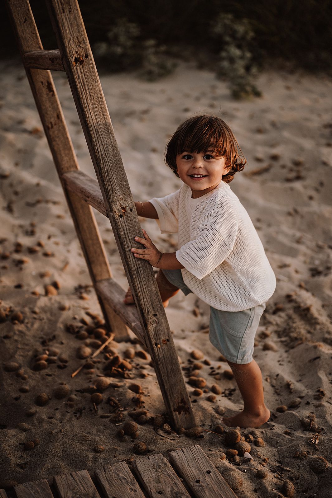 Smiling toddler climbing a wooden ladder on a sandy beach at golden hour.