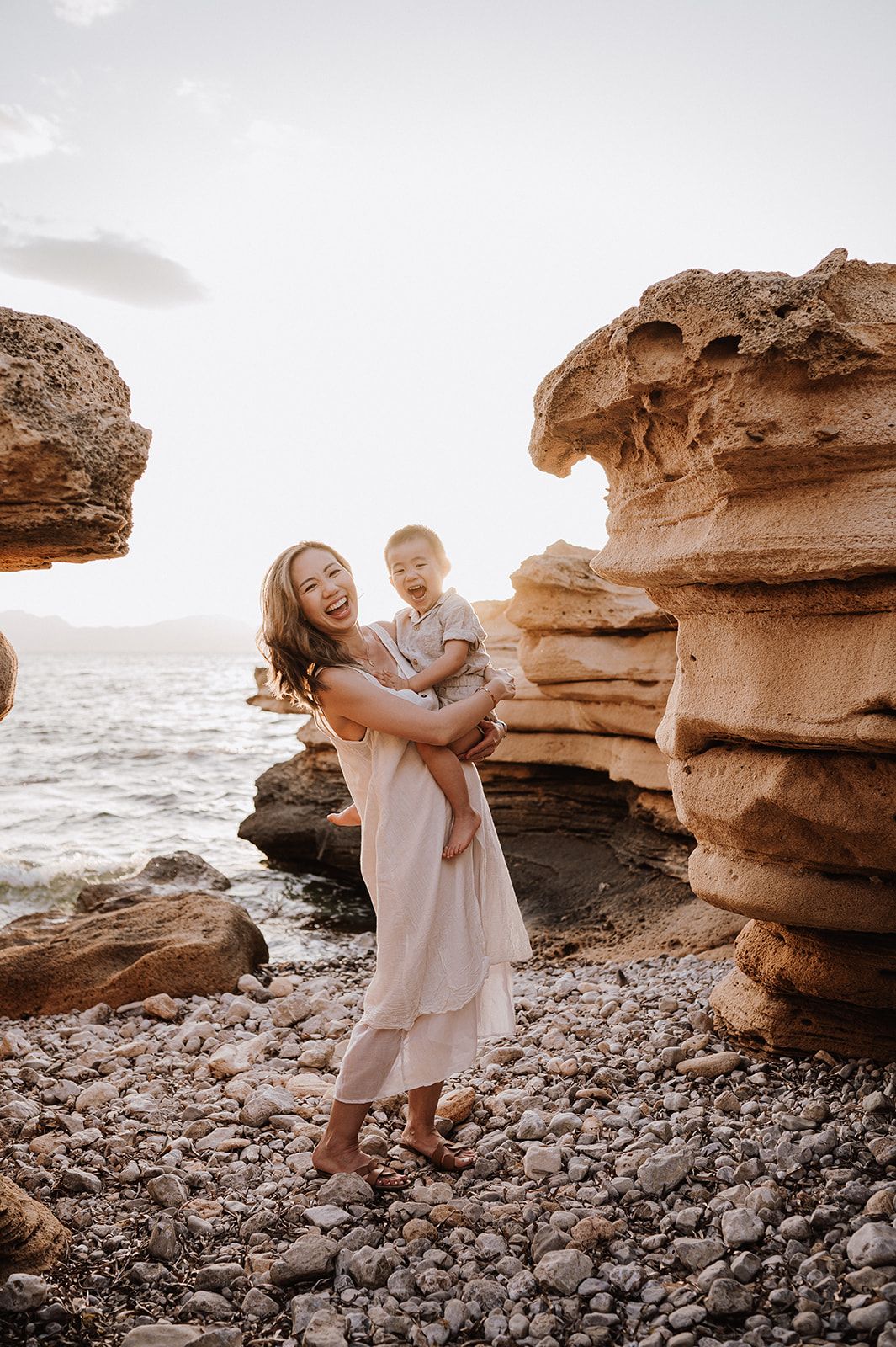 A joyful mother holds her laughing toddler on a rocky beach with golden sandstone formations.
