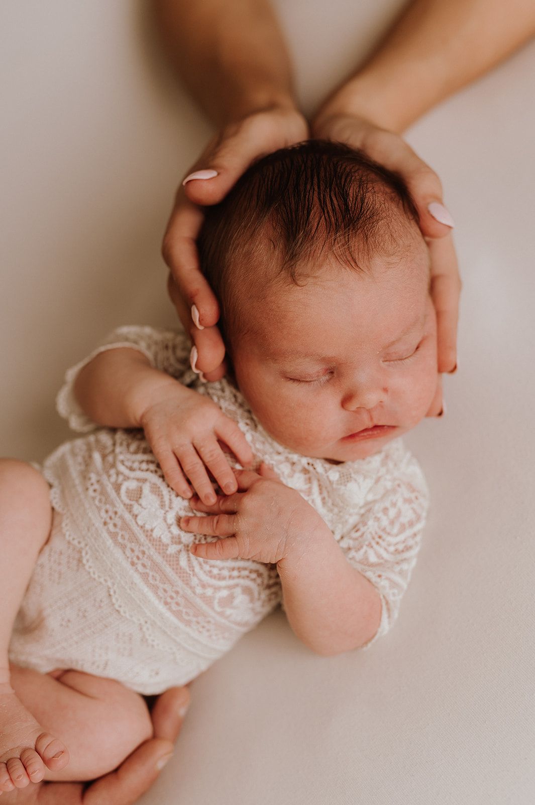 Newborn baby in white lace outfit sleeping peacefully while a parent gently cradles their head with both hands.