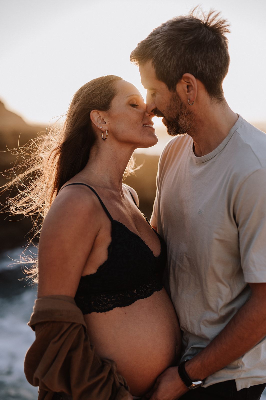 Pregnant couple sharing romantic moment at golden hour on rocky beach at sunset.