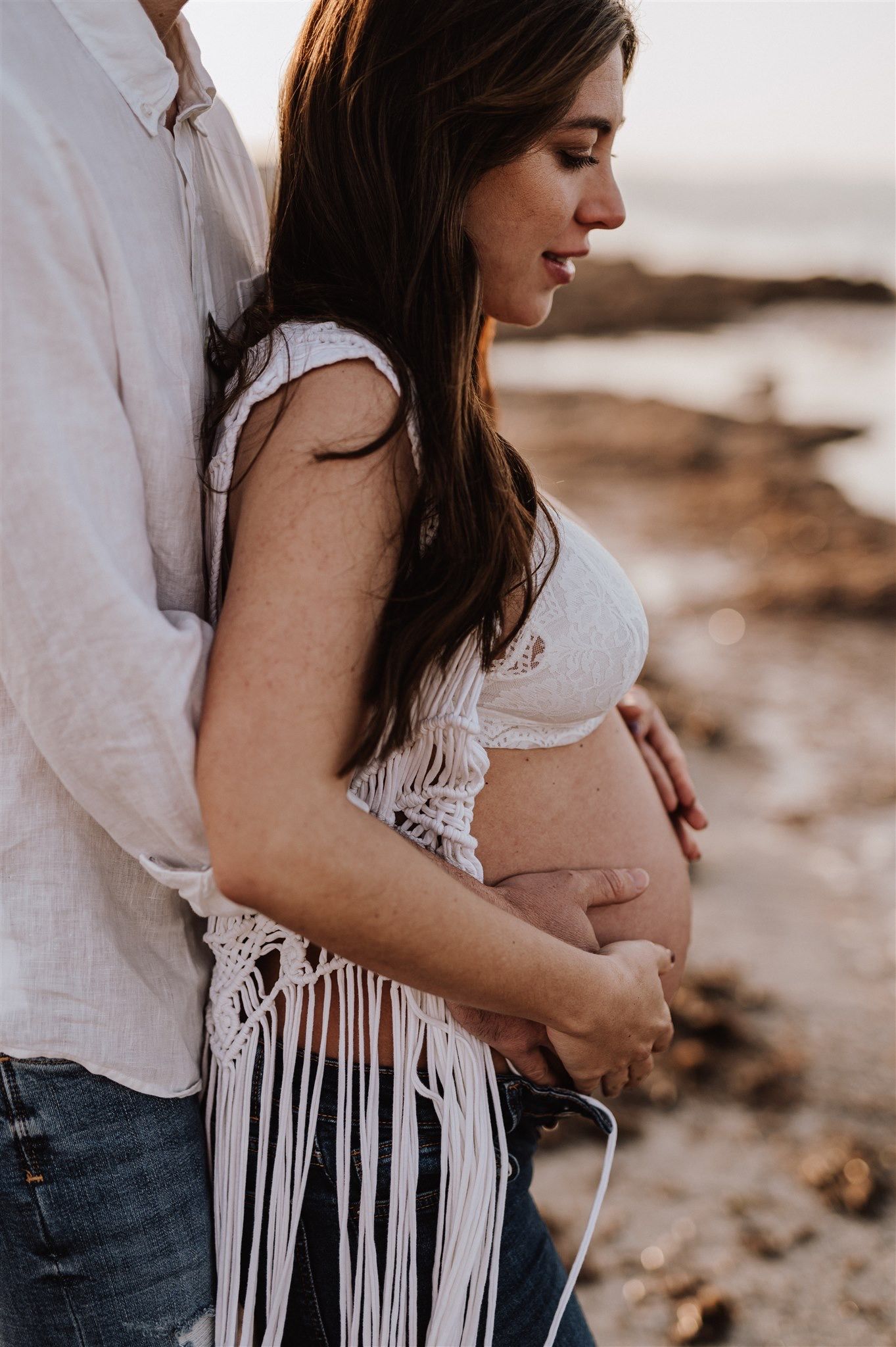 Pregnant woman in white macrame top holds her baby bump with partner embracing her on a rocky beach at sunset.