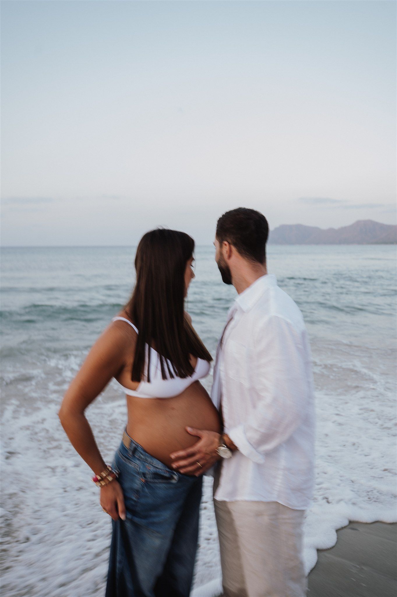 Pregnant couple standing on a beach at dusk, man gently touching woman's baby bump near ocean waves.