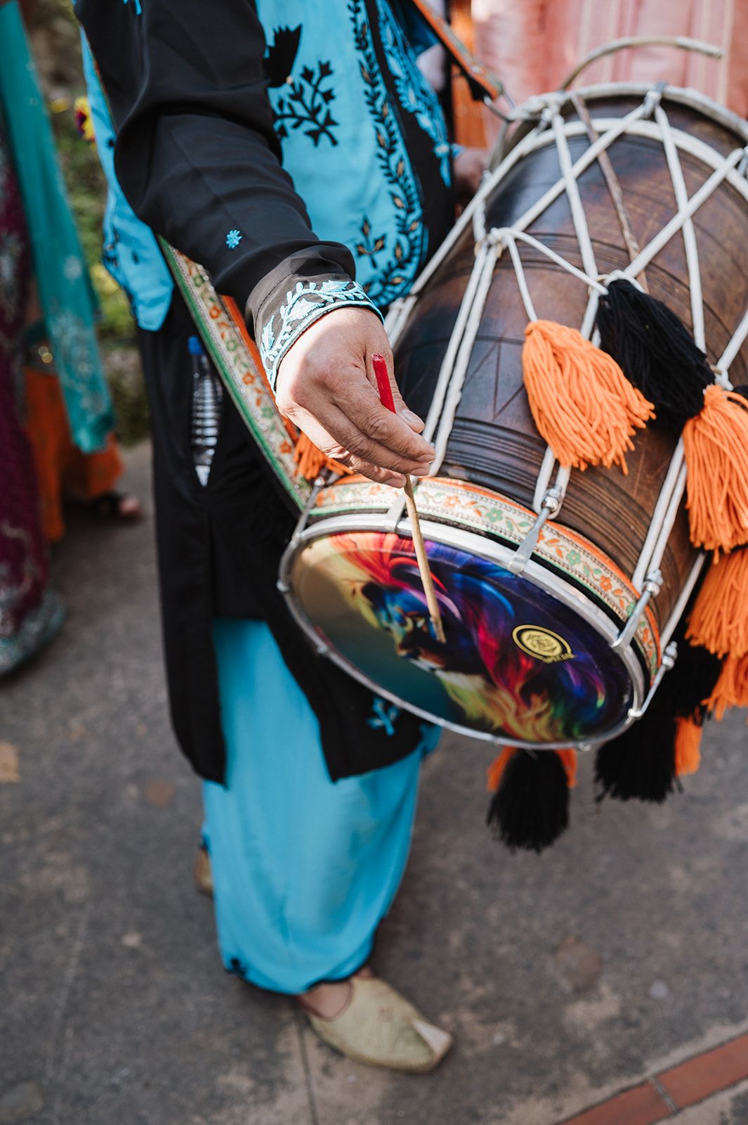 Close-up of hands playing a traditional dhol drum with orange tassels at an Indian wedding.