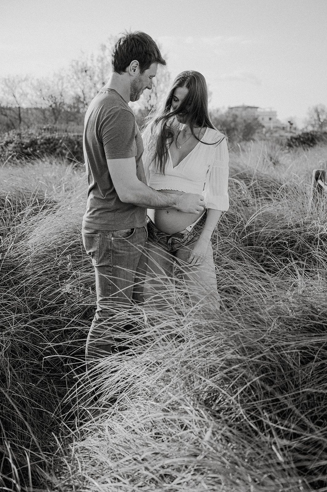 Black and white maternity photo of a couple embracing in tall grass field, woman showing baby bump.