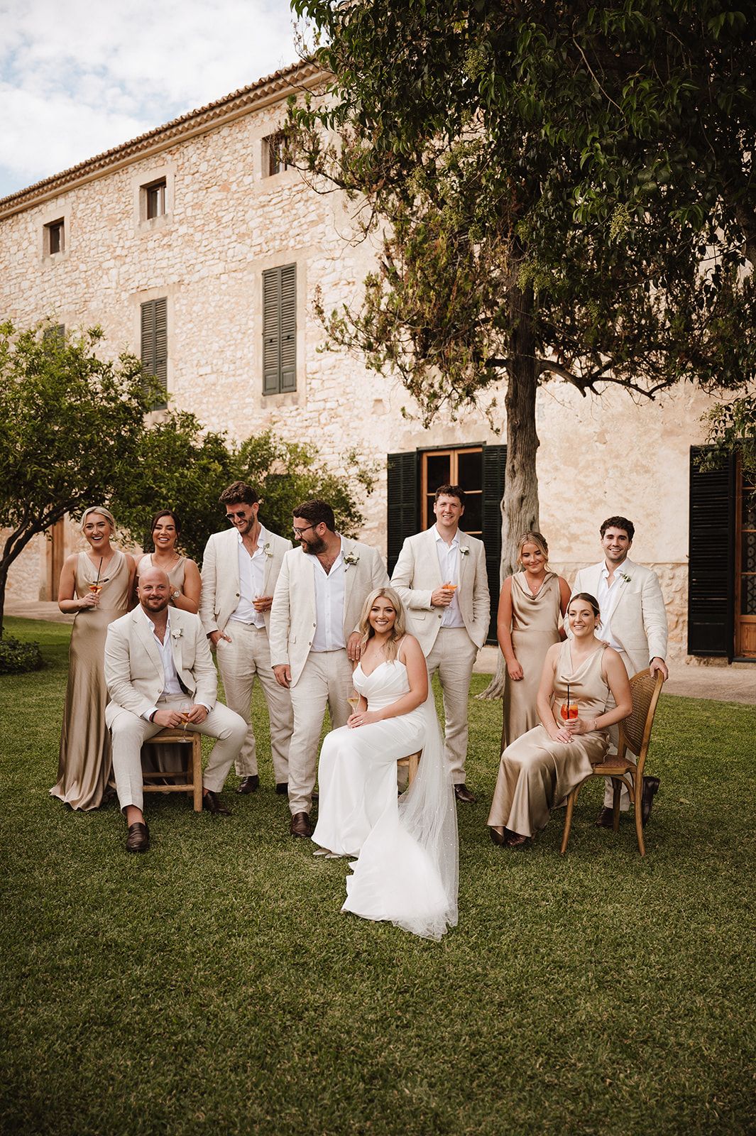 Wedding party posing casually on green lawn in front of a rustic stone building with bride seated center.