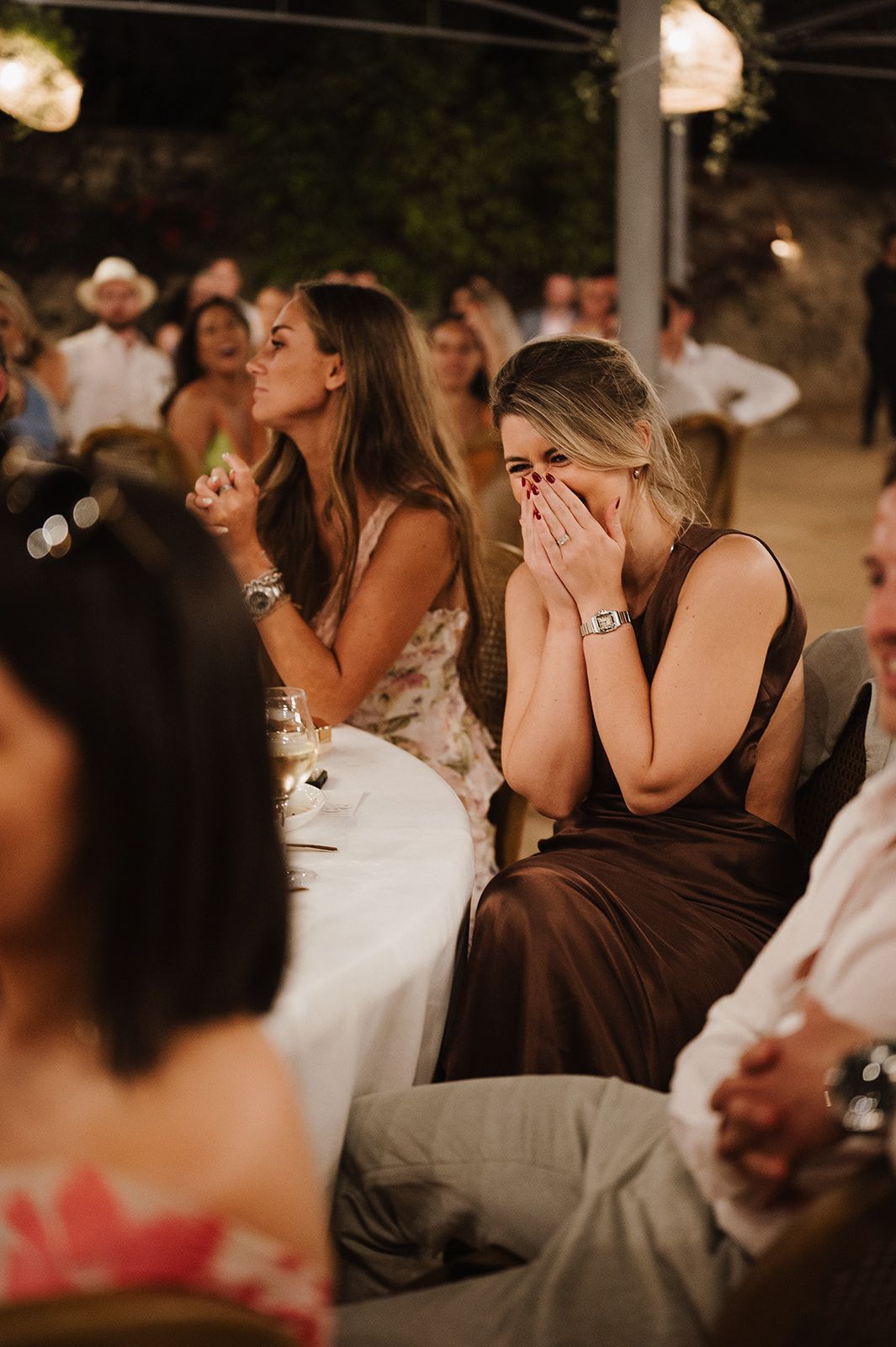 Emotional wedding guest covering mouth with hands during heartfelt speech at outdoor reception.
