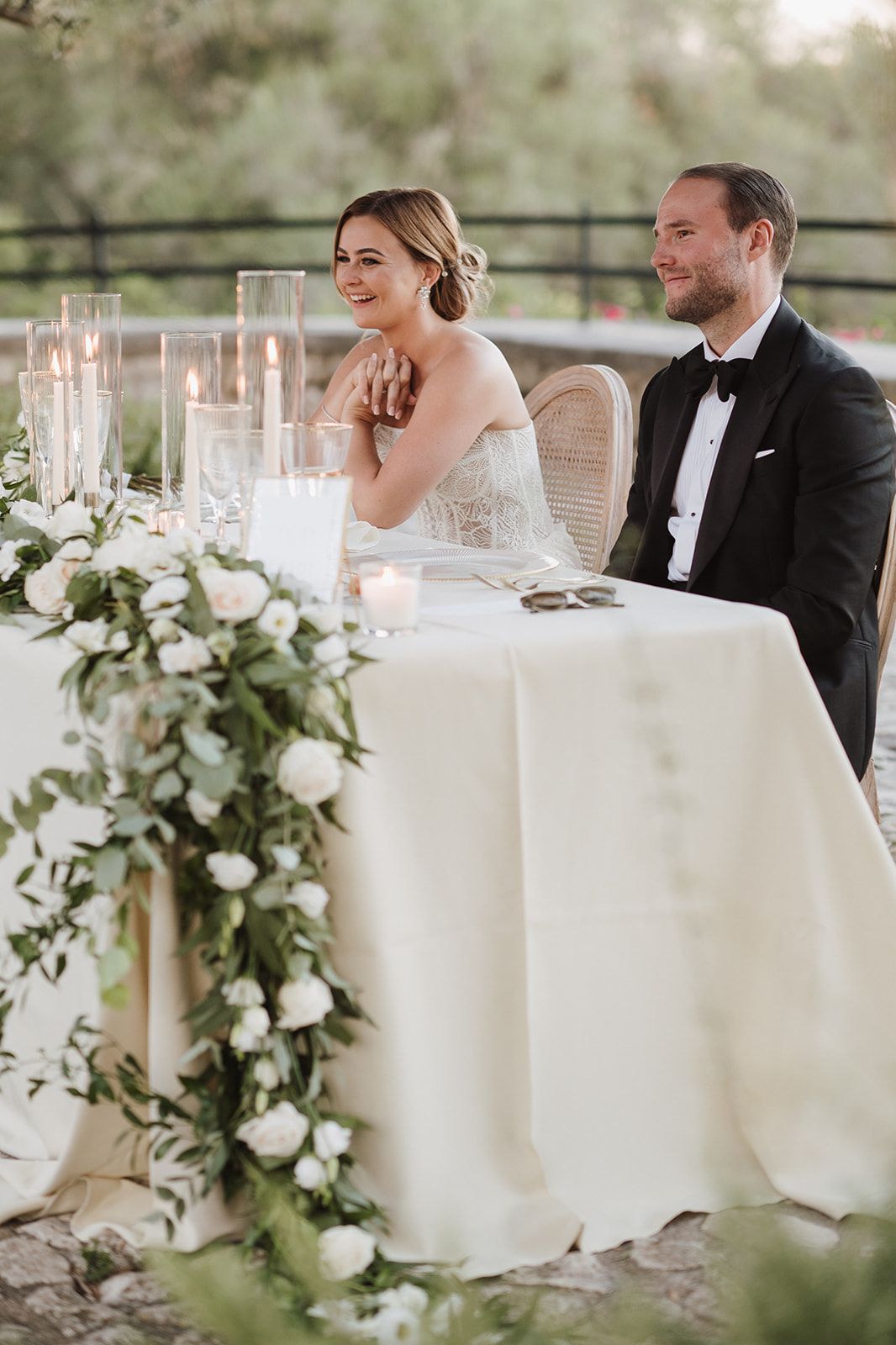 Bride and groom seated at sweetheart table adorned with white roses, greenery garland, and candles.