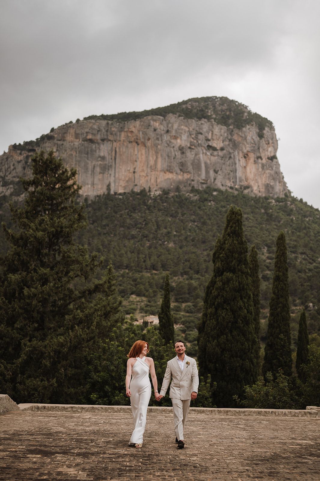 Bride and groom hold hands walking on stone terrace with dramatic rocky mountain backdrop.