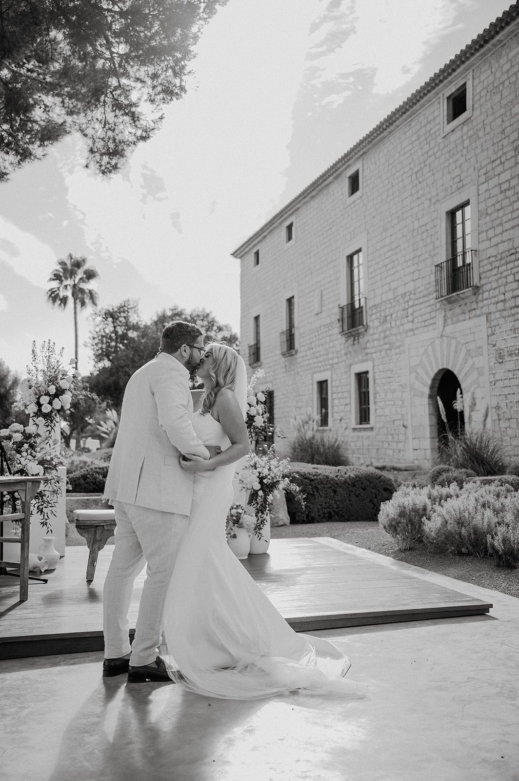 Bride and groom share first kiss at outdoor wedding ceremony beside a historic stone building.