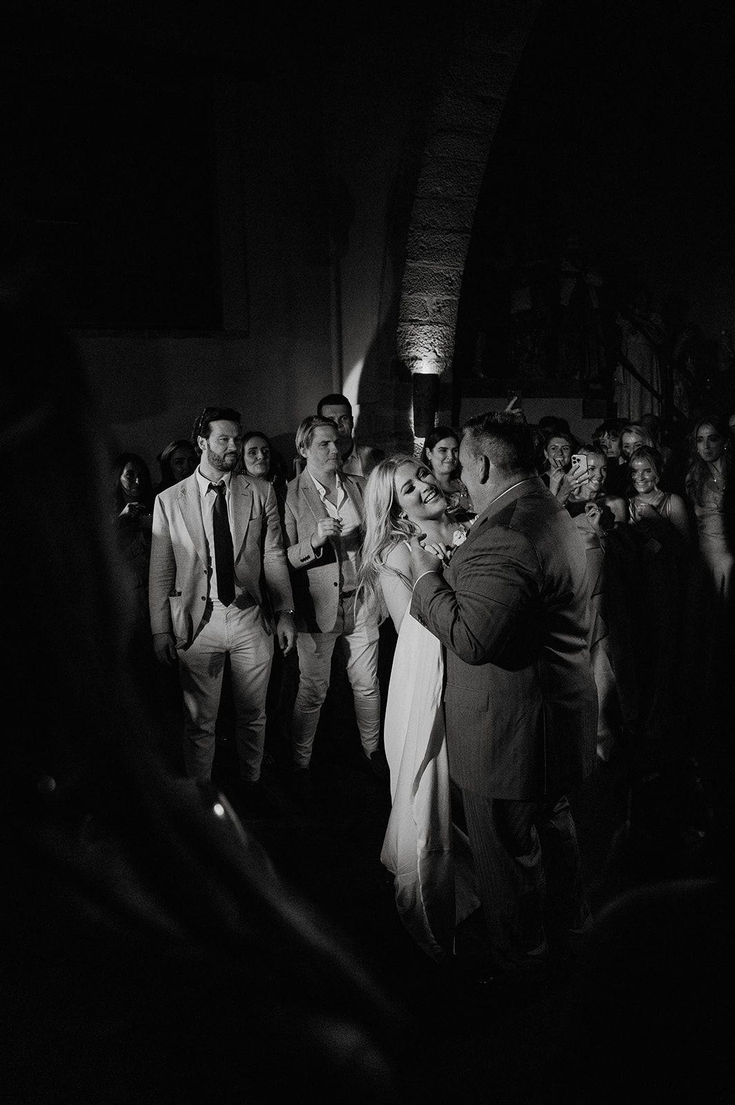 Black and white wedding photo of couple embracing on dance floor with guests watching.