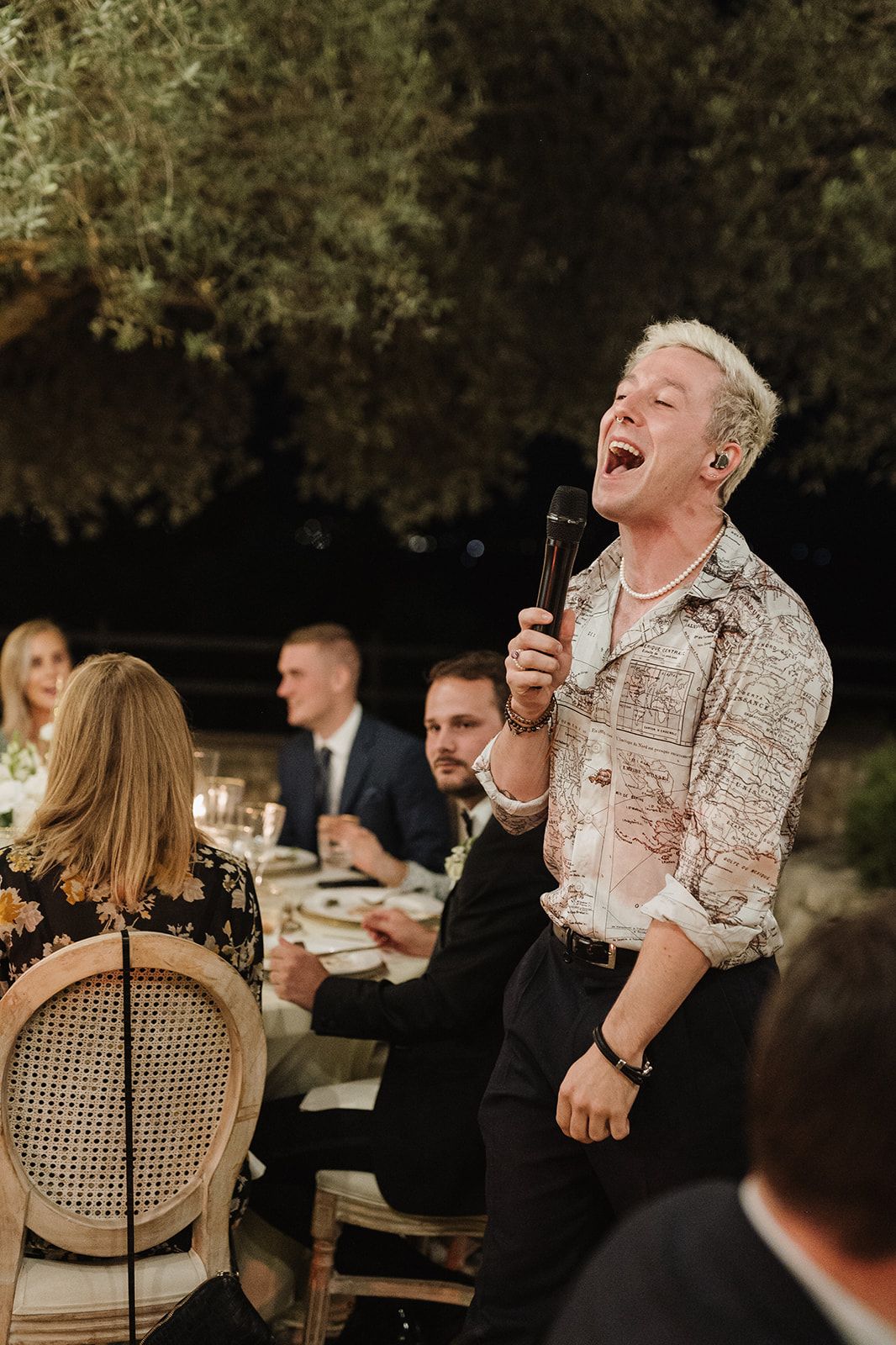 Blonde man in patterned jacket sings passionately into microphone at an outdoor wedding reception.