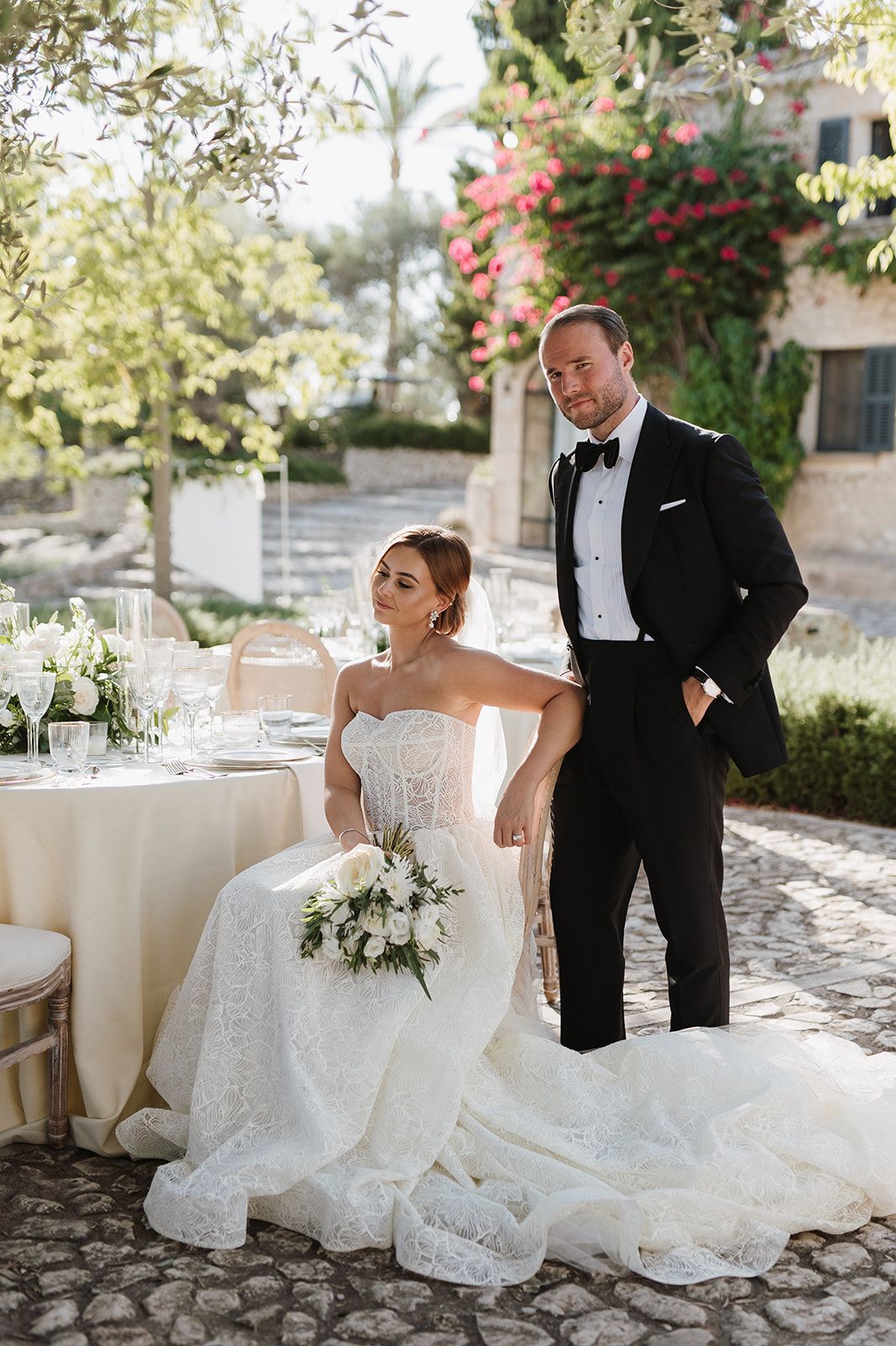 Elegant bride in lace gown seated at reception table with groom in black tuxedo in garden setting.