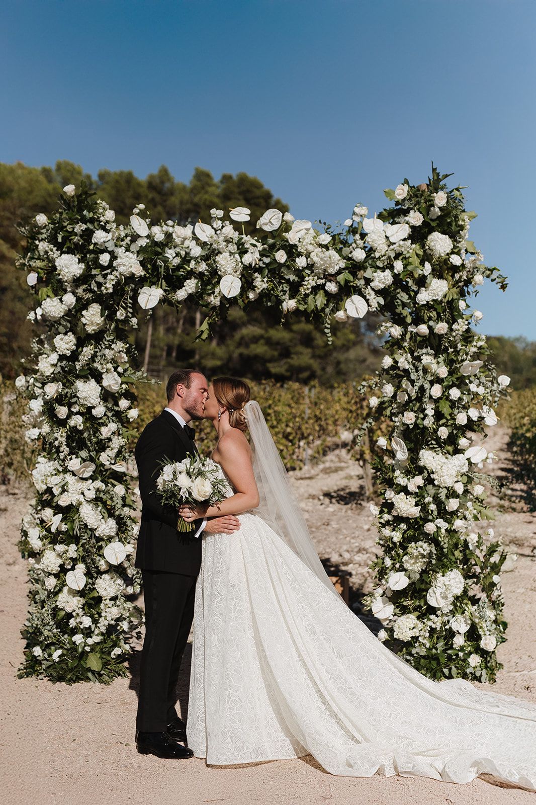Bride and groom share first kiss under white floral arch at vineyard wedding.
