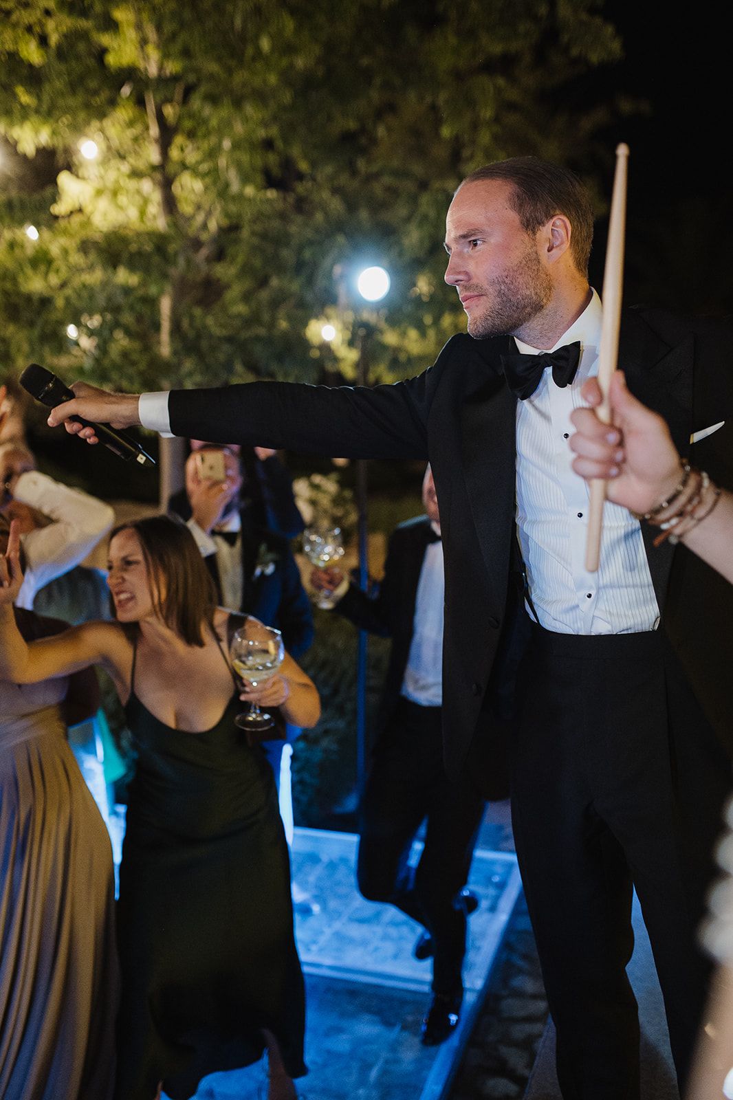 Tuxedo-clad wedding guest pours champagne while holding drumsticks during lively outdoor evening wedding celebration.