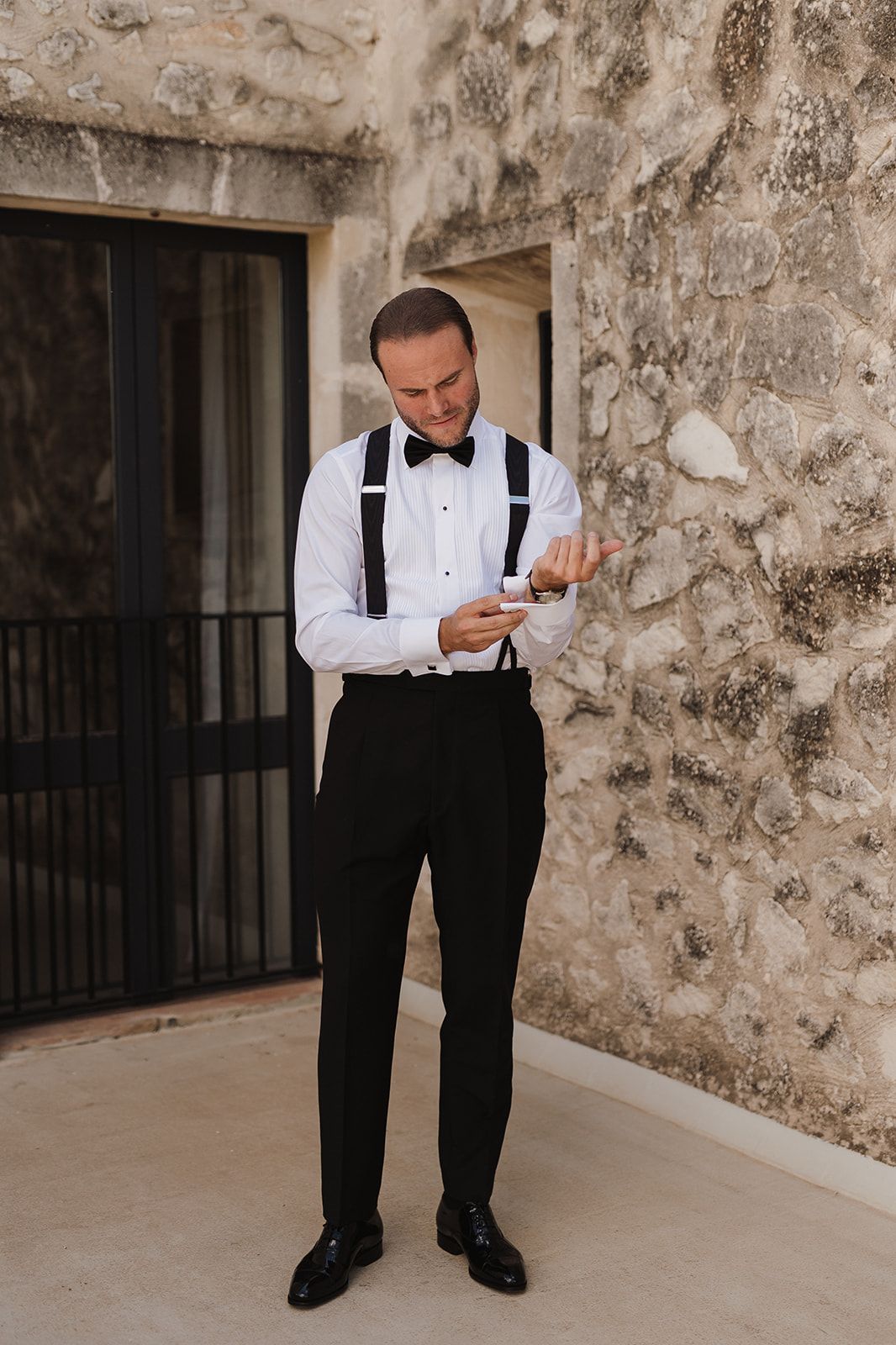 Dapper groom in black tuxedo with suspenders and bow tie checks his cufflinks against a rustic stone wall.