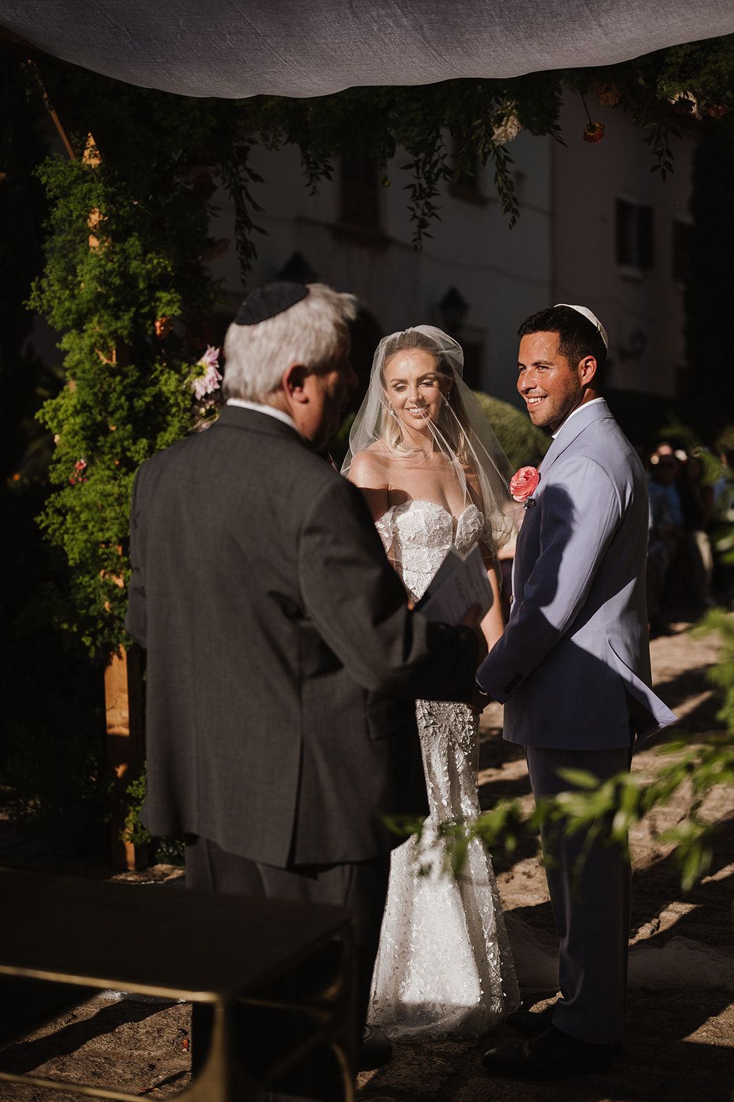 Bride and groom smiling during outdoor Jewish wedding ceremony with officiant under floral chuppah.