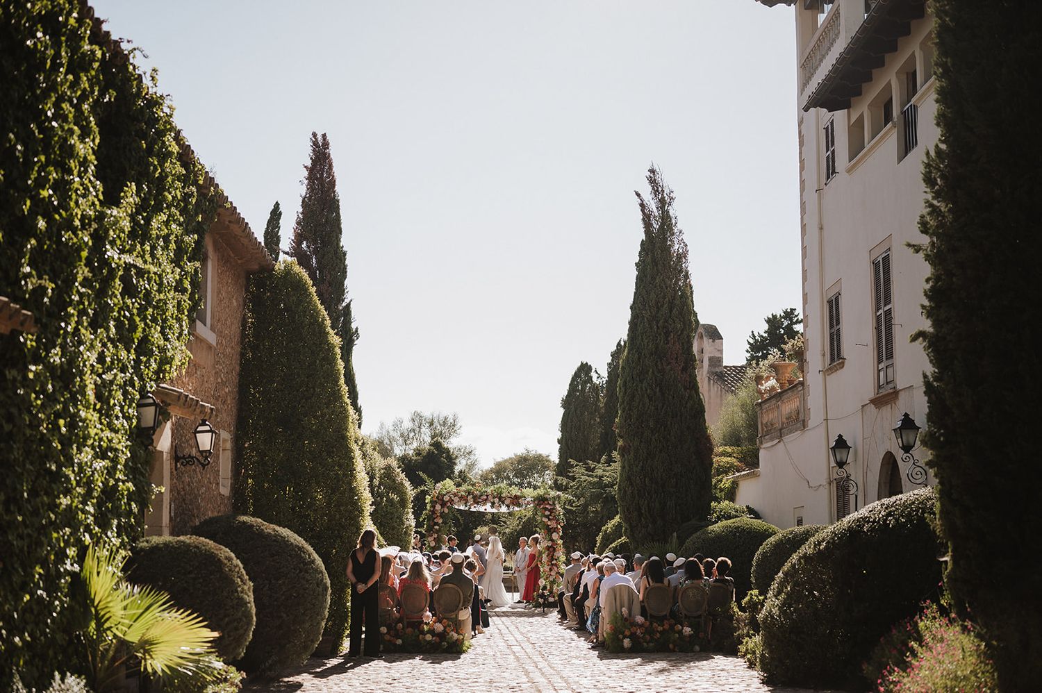 Outdoor wedding ceremony in a sunlit courtyard lined with cypress trees and manicured hedges.