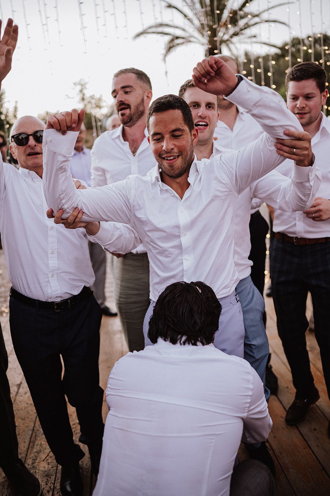 Groom celebrates joyfully with groomsmen in white shirts dancing under string lights at wedding reception.