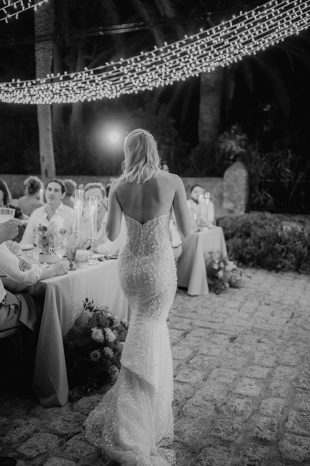 Bride in a stunning backless sequin gown walks through an outdoor reception lit by fairy lights.