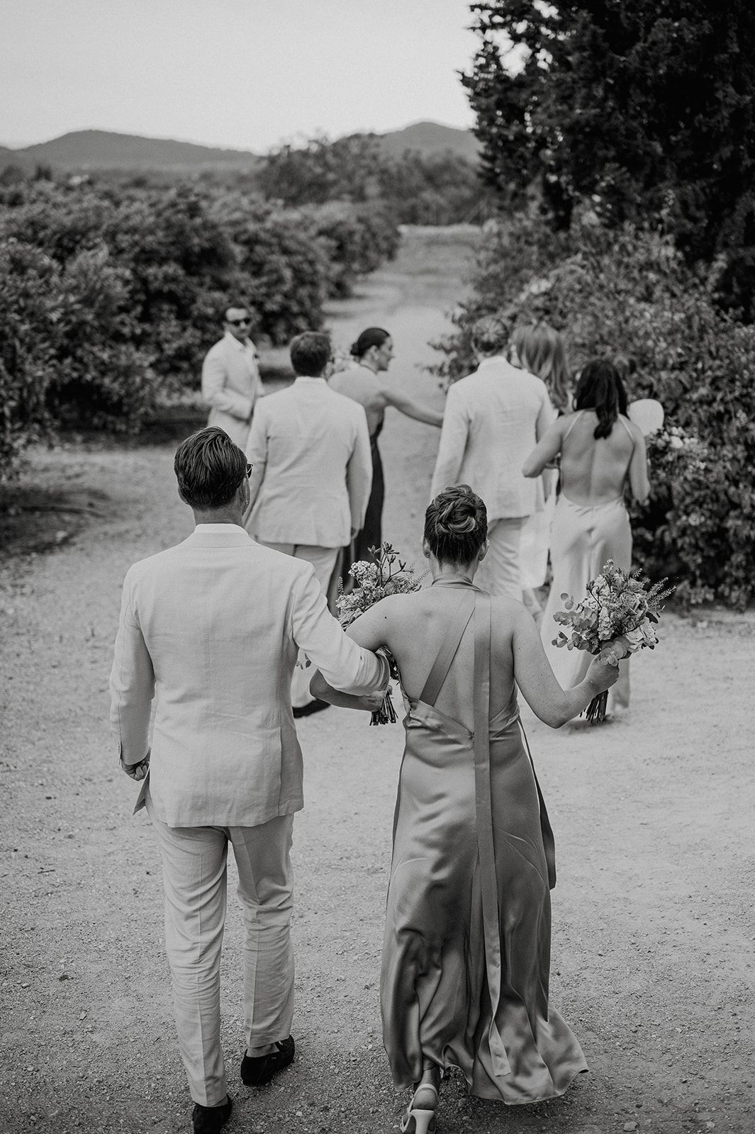 Black and white photo of wedding party walking along tree-lined path after outdoor ceremony.