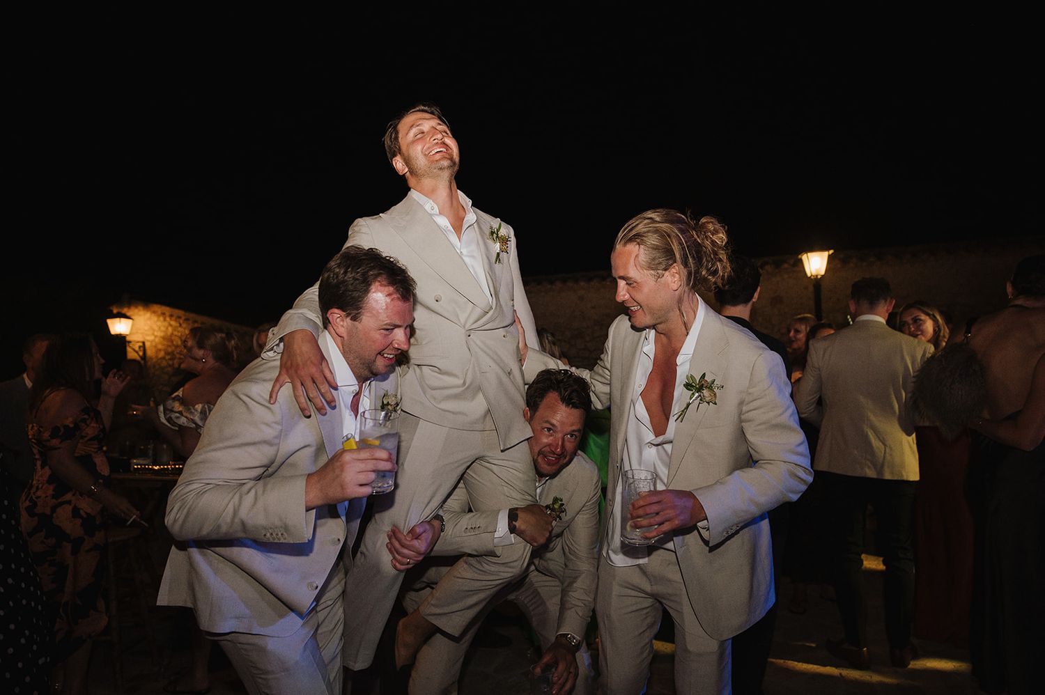Groomsmen in beige suits playfully lifting groom at an outdoor evening wedding reception.