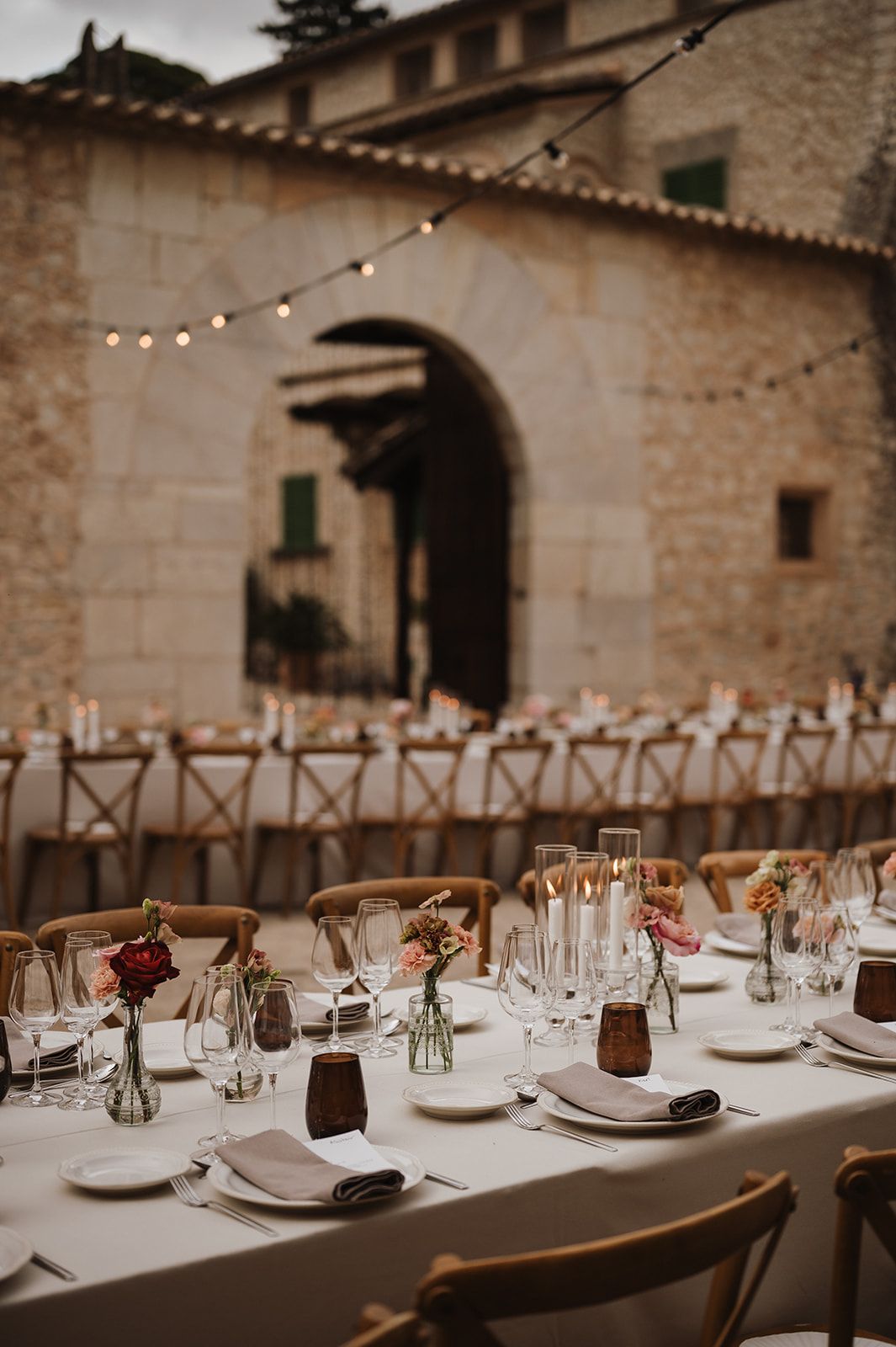 Elegant outdoor wedding reception with white tablecloths, bud vase florals, string lights, and stone arch backdrop.