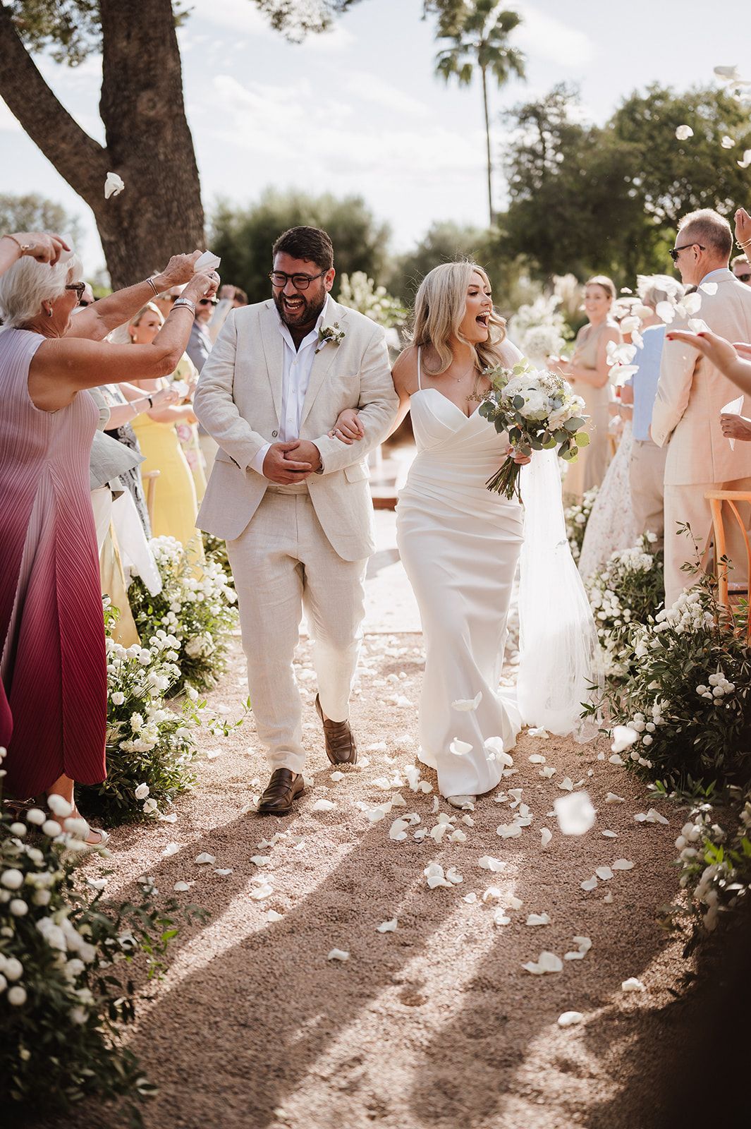 Joyful newlyweds walk down petal-lined aisle after outdoor wedding ceremony, guests tossing flowers in celebration.