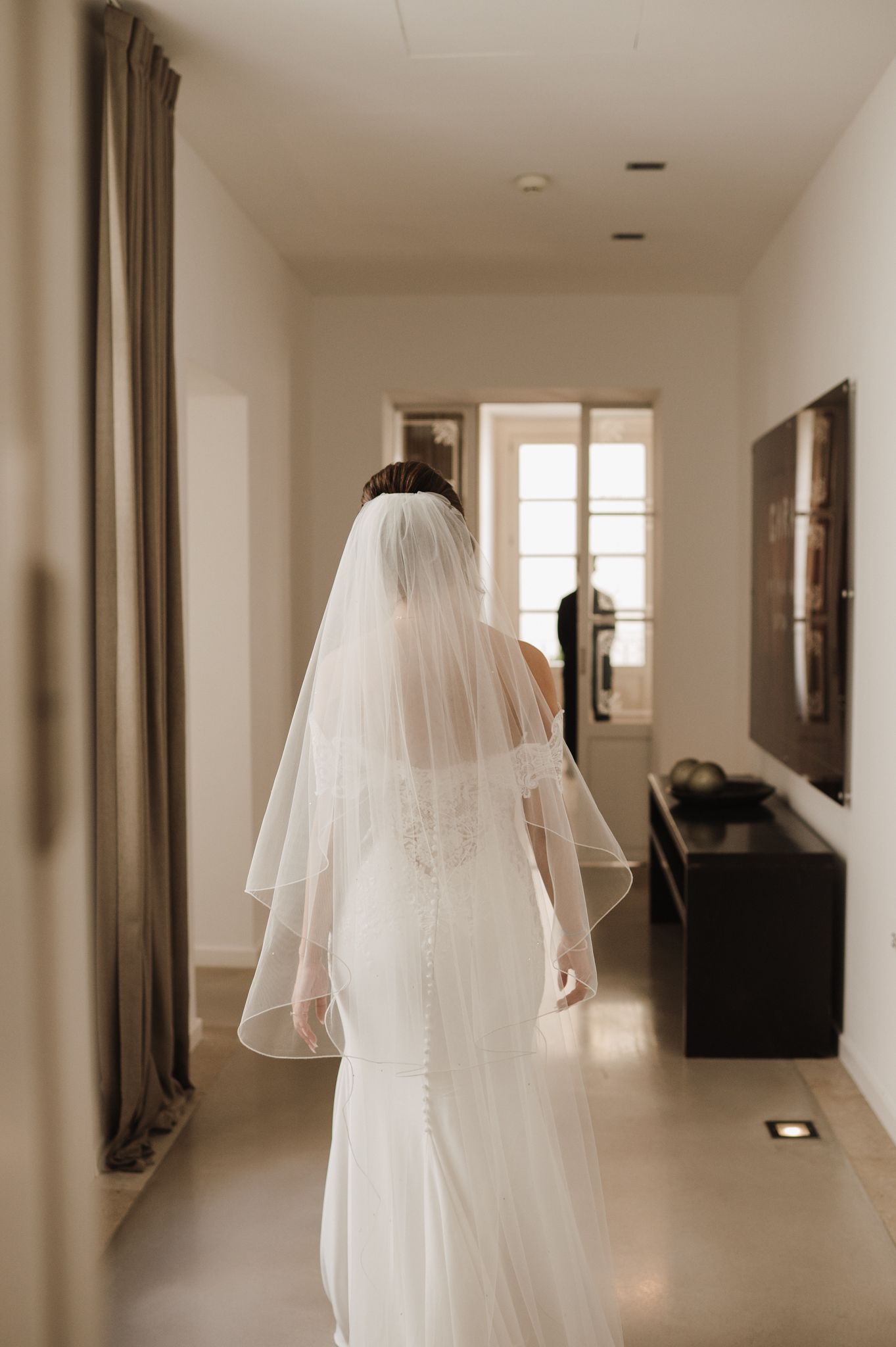 Bride in elegant white lace wedding gown and veil walks down hallway.