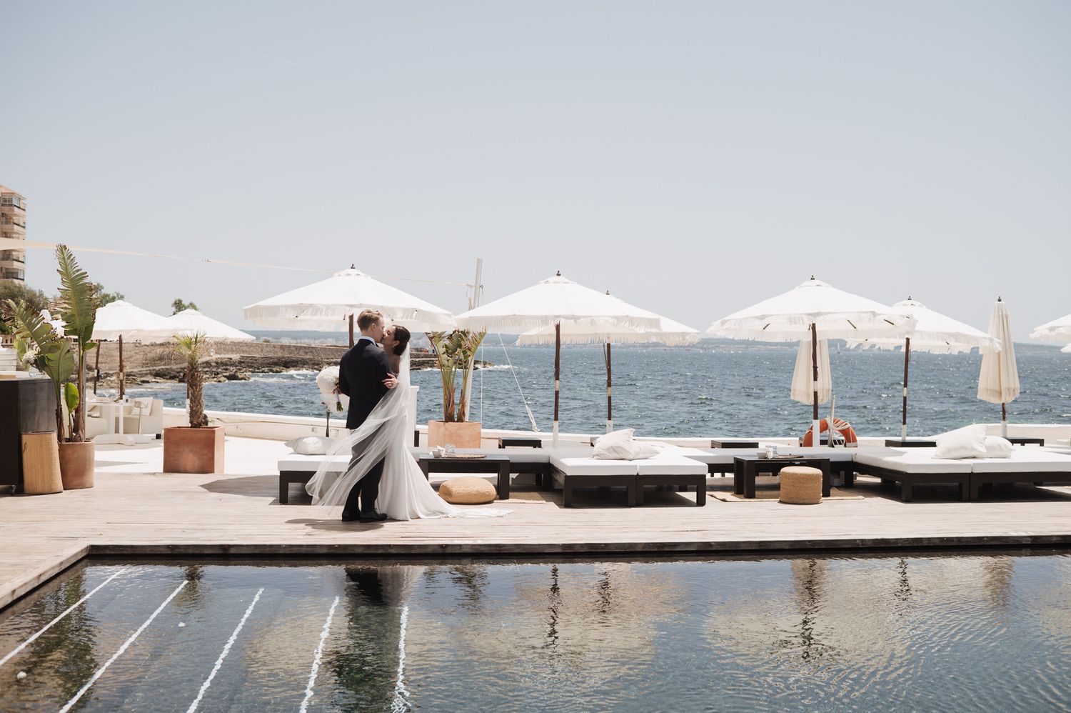 Bride and groom share a romantic dance beside a luxury pool overlooking the ocean at a seaside wedding venue.