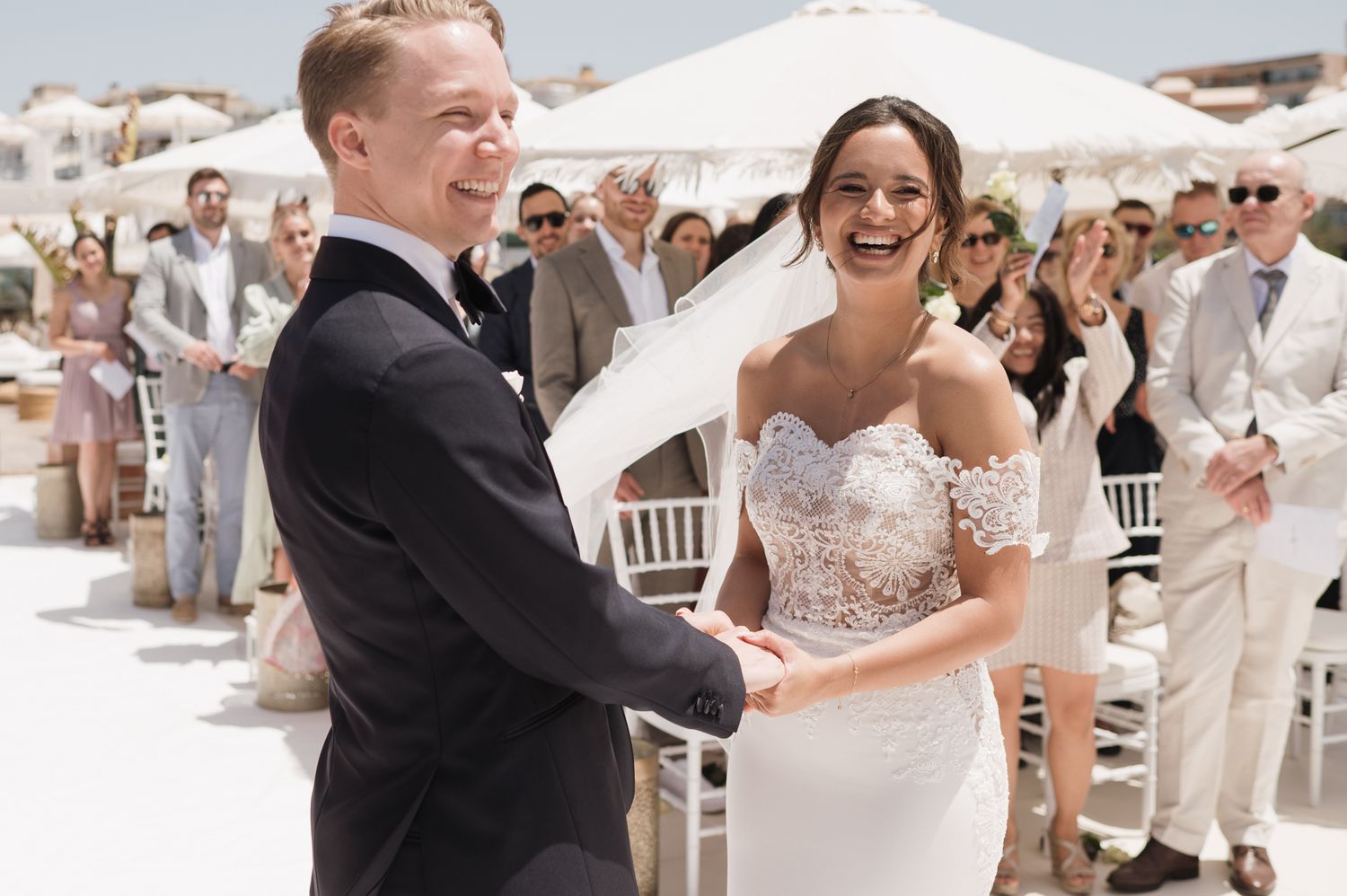 Joyful bride in lace gown and groom in tuxedo laugh during outdoor wedding ceremony with guests.