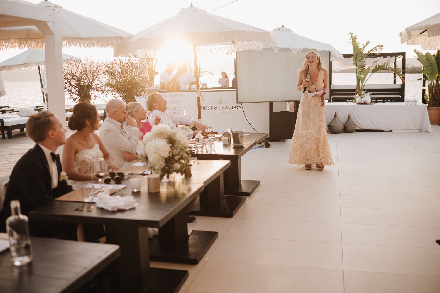 Blonde woman in blush gown delivers wedding speech to seated guests at outdoor sunset reception.