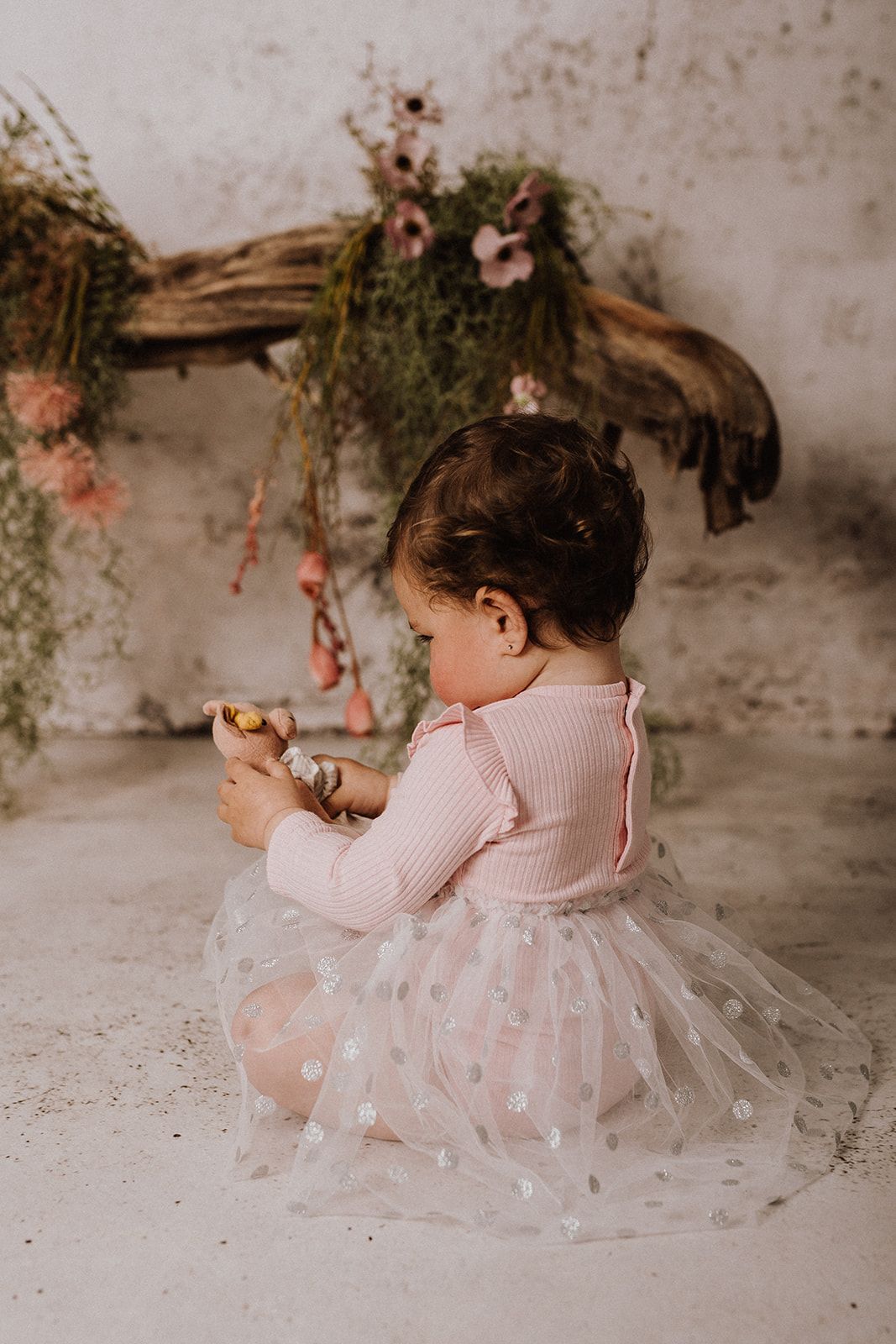 Baby girl in pink ribbed bodysuit and tulle skirt sits on floor holding toy near rustic floral decor.