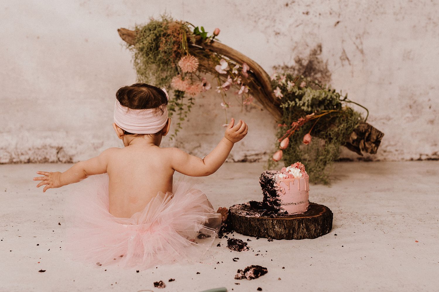 Baby girl in pink tutu smashes first birthday cake during professional photo session with floral arch backdrop.