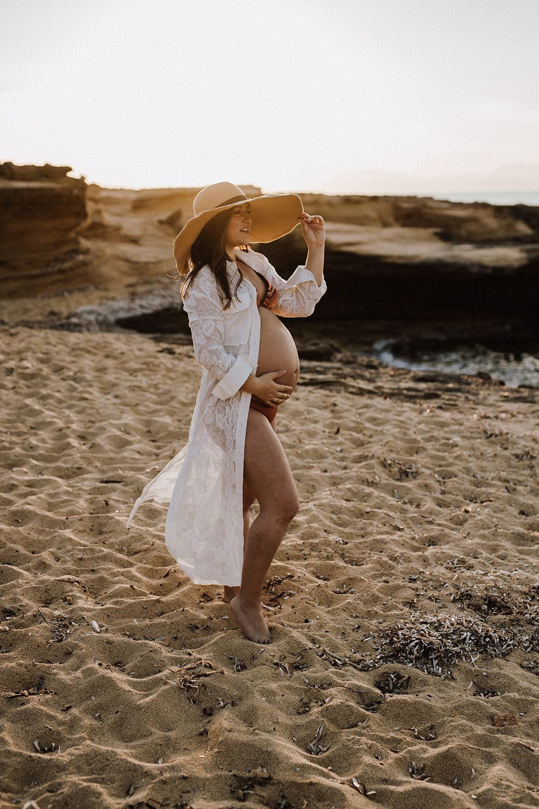 Pregnant woman in white lace dress and wide-brim hat cradling baby bump on sandy beach at golden sunset.