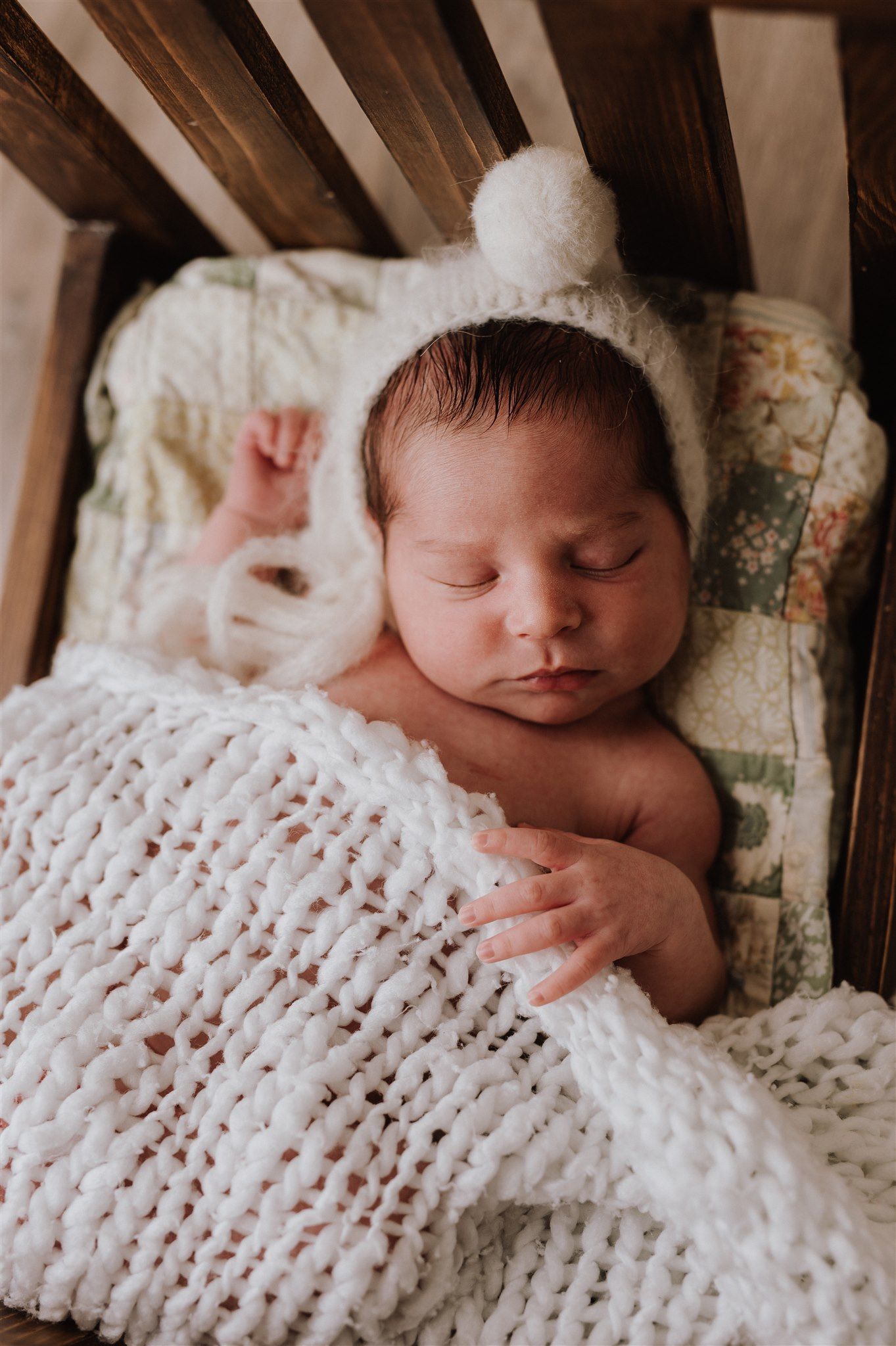 Sleeping newborn baby wearing a white knit bonnet with pompom, wrapped in a chunky knit blanket in a wooden crib.